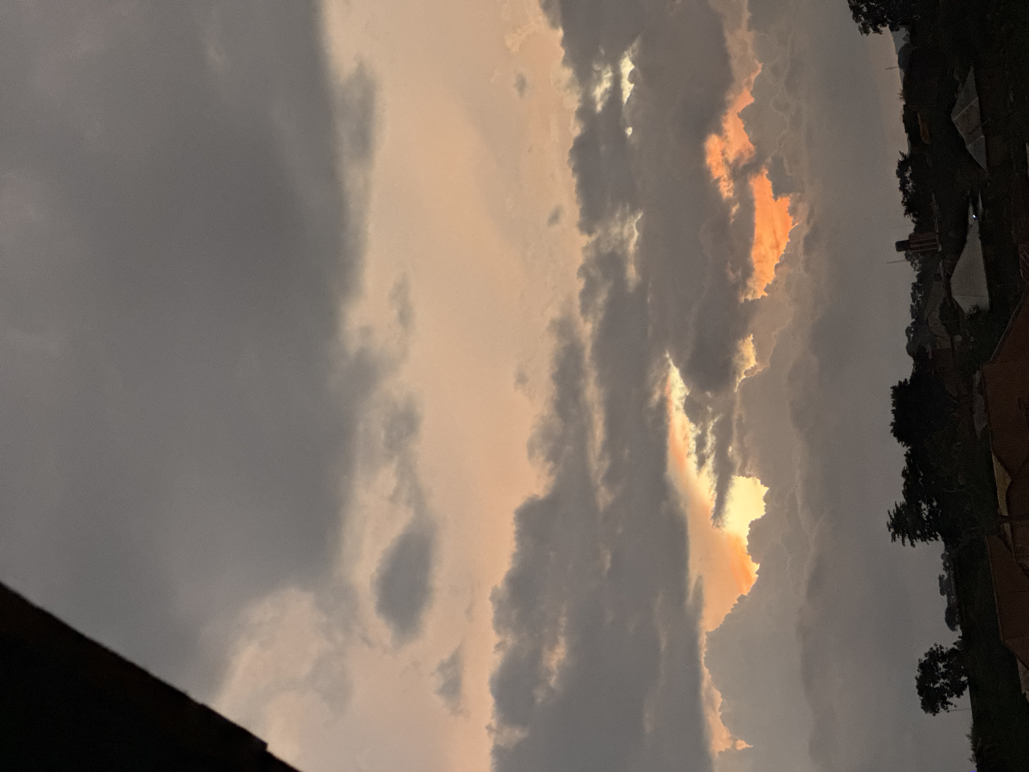 The image depicts a dramatic sunset sky over a city. Heavy, dark clouds dominate the upper portion of the frame, tinged with a soft orange glow from the setting sun. Breaks in the clouds reveal patches of vibrant orange and yellow light. Below the sky, silhouetted against the light, is the vague outline of a city or town, with buildings and trees visible. The overall impression is one of a moody and colorful atmospheric event.