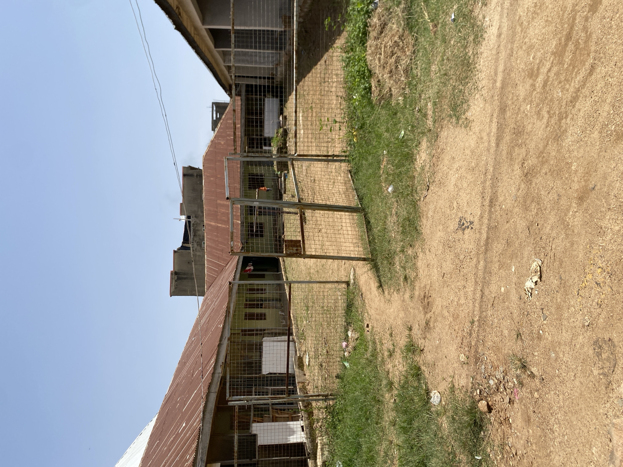 The image depicts an outdoor scene in Jos, Nigeria, under a clear blue sky. In the foreground, a dirt path leads towards a fenced-off area. The fence is made of wire mesh with metal posts, and a gate is partially open. To the left of the path and in front of the fence, there is overgrown grass and some scattered debris, including plastic bags and stones.
Beyond the fence, a courtyard or open space is visible. This area is also covered with dirt and sparse vegetation. In the background, there are several single-story buildings with rusty, corrugated metal roofs. The buildings appear to be constructed from light-colored materials, possibly concrete blocks, and have numerous windows, many of which are covered with wire mesh or bars. Some of the buildings have signs or white boards attached to their exteriors.
Further in the background, taller, more modern structures are visible, including a concrete building with a flat roof. Overhead, power lines stretch across the frame. The overall impression is one of a functional but somewhat rundown urban or semi-urban environment. There are no people visible in the image, and the scene appears to be one of an abandoned or less-maintained area.
