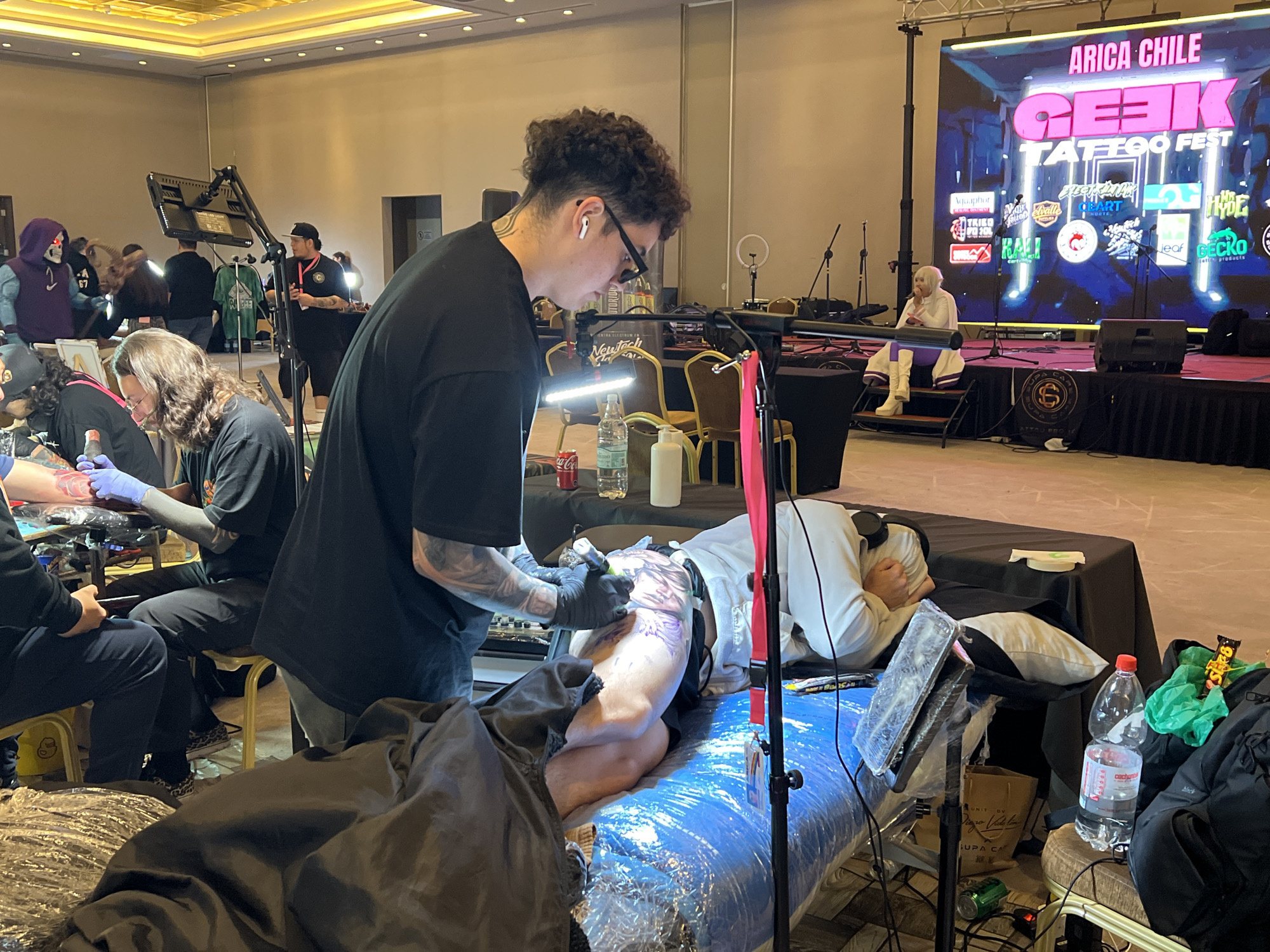 The image depicts a scene at the "Arica Chile Geek Tattoo Fest" held in a large, well-lit indoor venue, possibly a convention center or ballroom, characterized by a high, illuminated ceiling. The focus is on a tattoo artist, a young male with curly hair and glasses, wearing a black t-shirt and earbuds, actively tattooing a client. The client, lying on a tattoo bed covered with blue plastic, is a person with light skin and is wearing a white shirt. The artist is wearing black gloves and holding a tattoo gun.
Several other individuals are present in the background. To the left, a person in a purple hooded costume with a skull mask is visible. Further back, other attendees are seen standing and observing. A stage is set up in the background with a large LED screen displaying the event's name, "ARICA CHILE GEEK TATTOO FEST," along with various sponsor logos. There are microphones and speakers on the stage, suggesting performances or presentations are part of the event.
On a table near the tattoo station, there are bottles of water, a can of soda, and other supplies. The overall environment is consistent with an organized event, with defined areas for tattooing and a backdrop indicating a public gathering. The lighting suggests an indoor setting with artificial illumination.