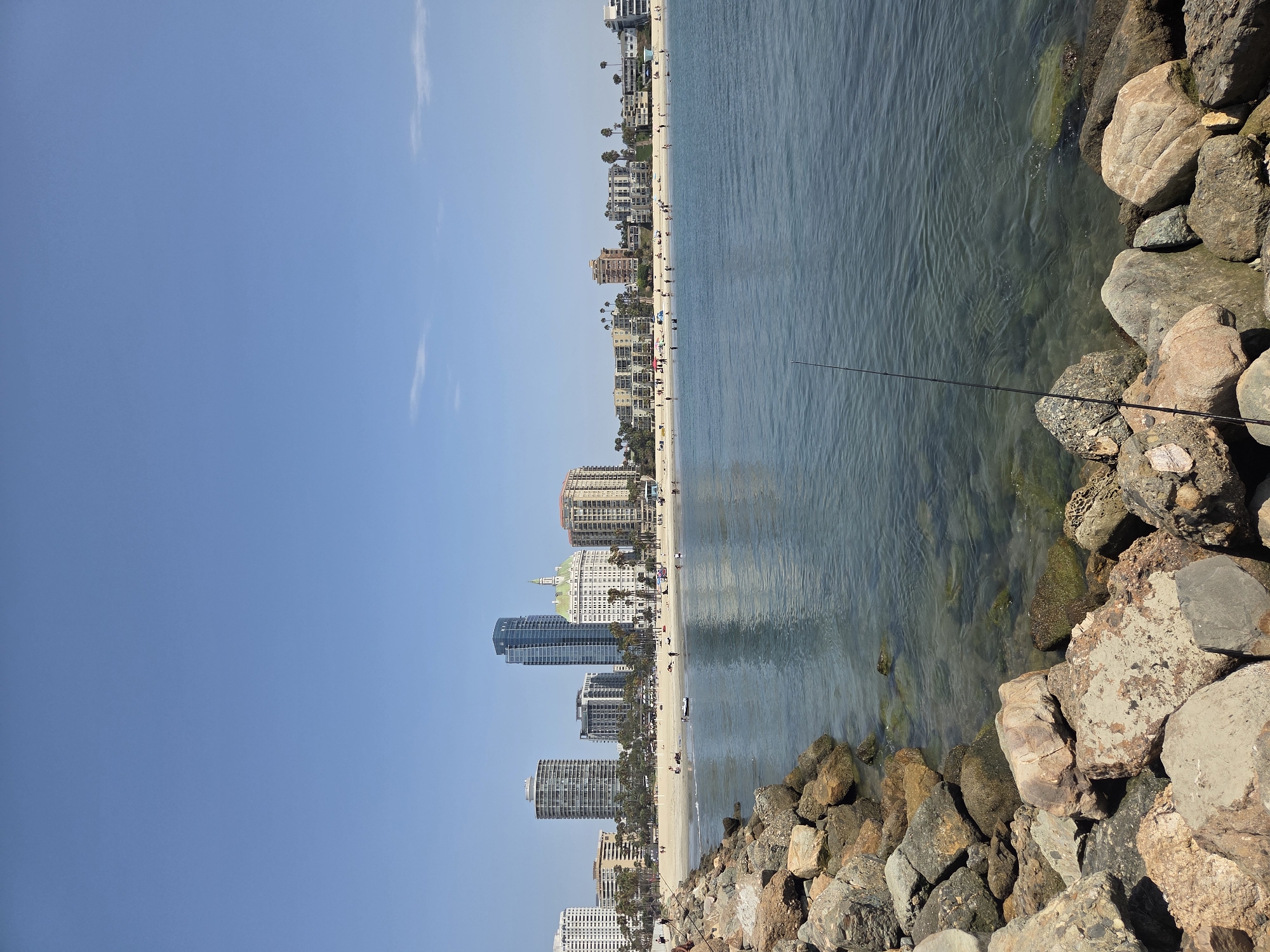 The image depicts a coastal scene in Long Beach, California, United States of America. In the foreground, a rocky breakwater is visible, with large, weathered stones leading into the clear, blue water. The water itself appears calm, with ripples on the surface and a visible seabed in shallower areas.
Beyond the water, a sandy beach stretches across the mid-ground. Numerous people are spread across the beach, some sitting or lying down, while others are walking or wading in the shallows. Beach umbrellas and tents dot the sand, suggesting a leisurely day.
The background is dominated by the Long Beach skyline, featuring a variety of buildings. Tall, modern high-rise buildings, including a prominent blue skyscraper, stand alongside lower-rise apartment complexes and hotels. Palm trees are interspersed among the buildings, indicating a warm climate. The sky above is a clear, bright blue with only a few wispy clouds on the horizon. A single fishing rod is planted in the rocks in the lower right quadrant of the image, extending into the water.