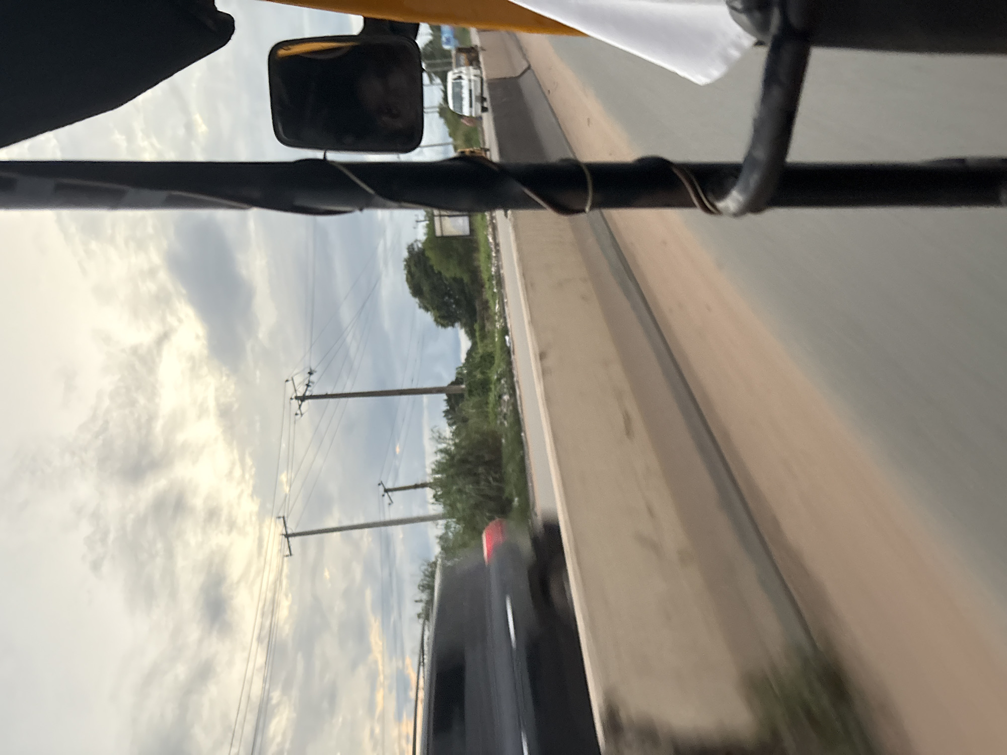 The image captures a view from the interior of a tuk-tuk, looking outwards onto a road in Onitsha, Nigeria. The sky above is overcast with grey clouds, suggesting a potentially humid or rainy day. In the foreground, a black metal pole, likely part of the tuk-tuk's structure, runs vertically through the frame, with a coiled wire wrapped around it. A side mirror is visible, reflecting a dark interior.
On the road, a dark-colored SUV is seen in motion, slightly blurred due to the movement of the tuk-tuk. The SUV's taillights are illuminated, indicating its presence on the road. Beyond the road, there is a barrier made of concrete blocks, separating the road from a grassy area. Several utility poles with wires stretch across the landscape, a common sight in urban and semi-urban areas. In the distance, a white van and another vehicle are also visible on the road. The environment appears somewhat unkempt, with vegetation and what looks like debris present along the roadside. The overall impression is a snapshot of everyday life and transportation in Onitsha.