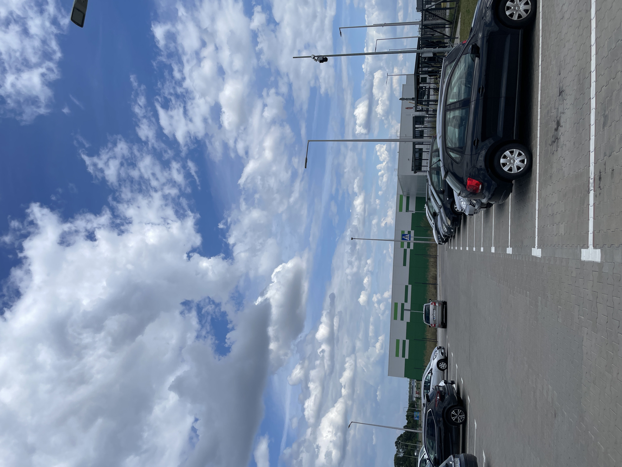 This is an outdoor, eye-level, medium shot of a parking lot in Nowa Wieś Kącka, Poland. The sky is filled with large, white, fluffy clouds against a bright blue backdrop. In the foreground, a black Volkswagen Polo is parked. To the left, other cars are visible, including a silver Skoda Fabia. In the background, a large building with a green and white striped facade is situated behind a row of parked cars. Several streetlights are positioned throughout the parking lot.
