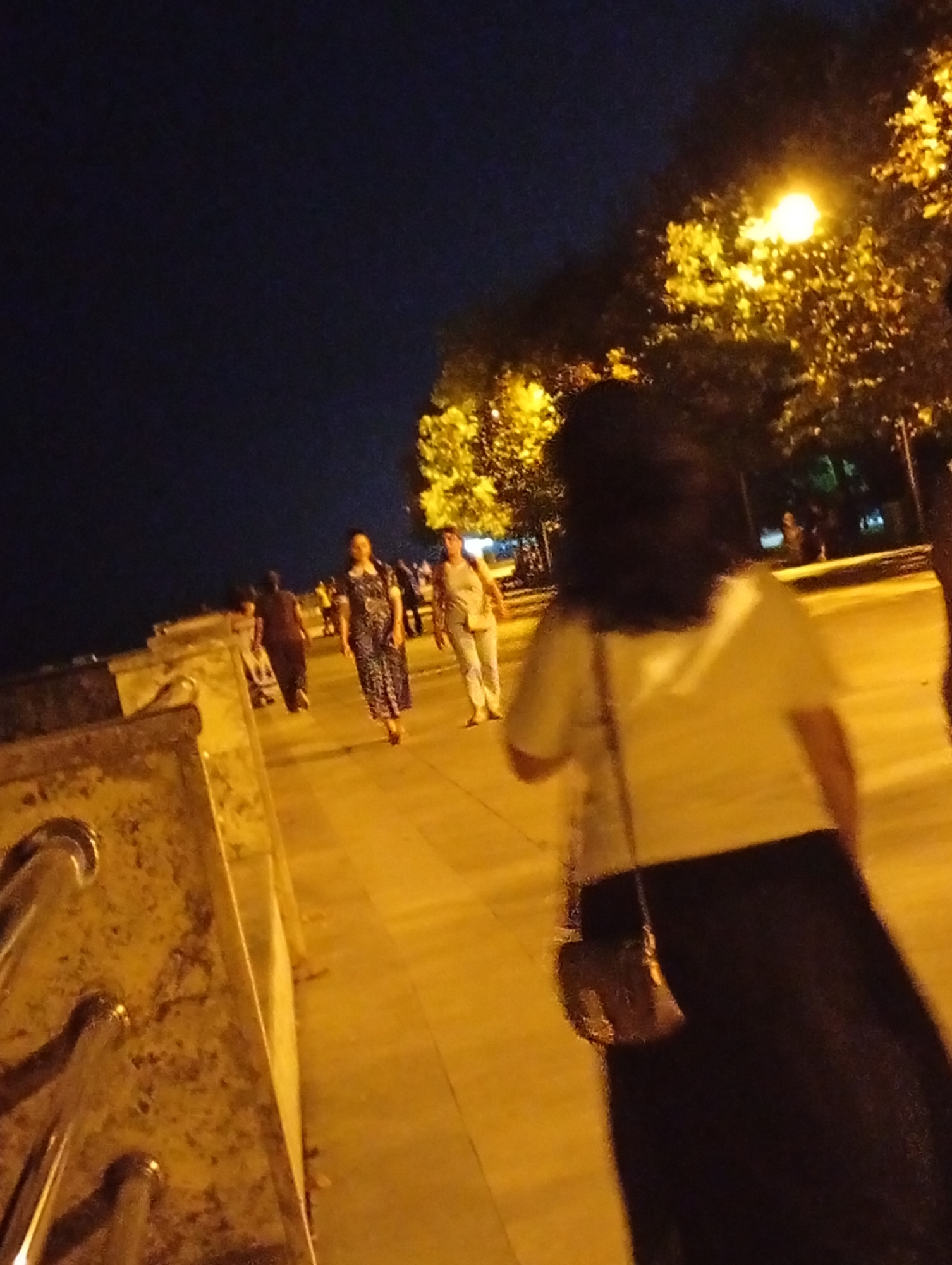The image depicts a nighttime scene along a waterfront promenade in Astara, Azerbaijan. Several individuals are walking along the illuminated path. In the foreground, a person with dark, long hair is visible from behind, wearing a light-colored top and a dark skirt. They are carrying a small, dark purse with a chain strap. To the left, a stone embankment with metal railings is prominent. Further down the promenade, a woman in a patterned dress is walking, and another person in light-colored clothing is ahead of her. Trees with yellow-tinged foliage, illuminated by streetlights, line the promenade, creating a warm glow against the dark night sky. The overall atmosphere is that of a public space bustling with evening activity.
