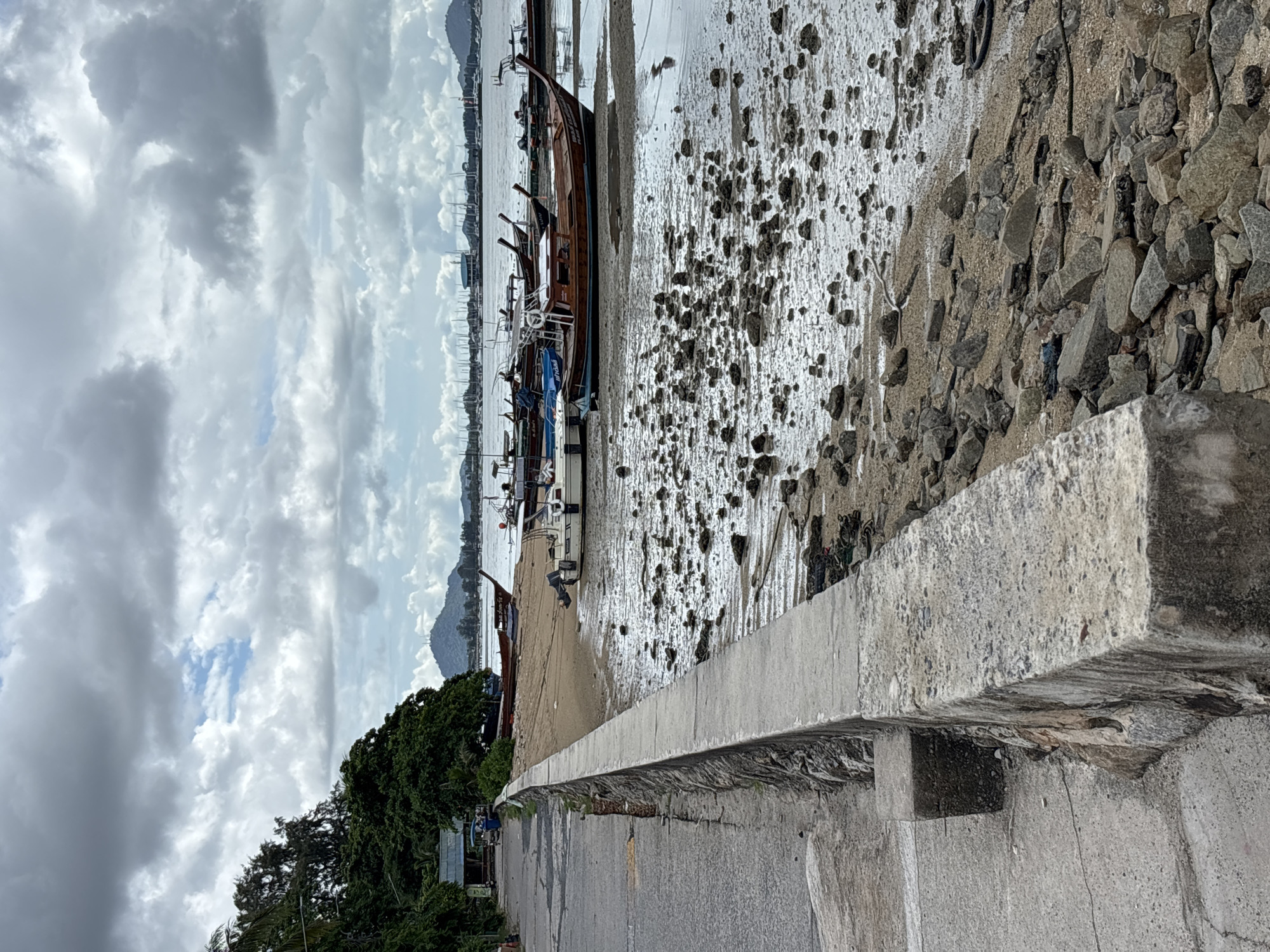 The image depicts a coastal scene in Rawai, Thailand, with a focus on the shoreline and moored boats. In the foreground, a concrete walkway runs along the left side of the frame, bordering a low stone wall. The wall separates the walkway from a sandy, rocky foreshore. The foreshore is covered with numerous stones and some debris, indicating it is likely low tide. Several boats, including traditional longtail boats and smaller motorboats, are grounded on the sand or partially submerged in shallow water. The water appears calm, reflecting the cloudy sky. In the background, a line of trees and vegetation separates the shore from a road and some low-rise buildings. Further in the distance, a hazy silhouette of hills can be seen against the overcast sky. The overall impression is one of a quiet, somewhat weathered coastal area. There are no people visible in the frame.