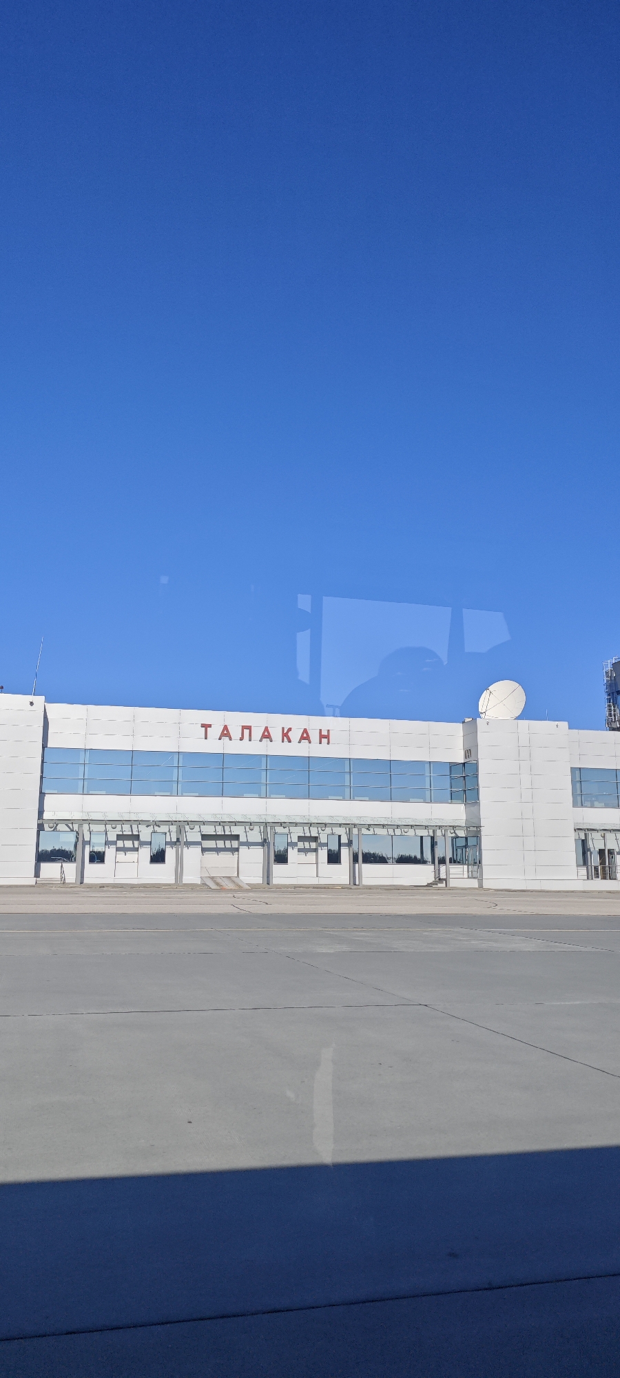 The image shows the exterior of the Talakan Airport building in Talakan, Russia on a clear, sunny day. The building is a modern, rectangular structure with a white facade, large windows, and the name "ТАЛАКАН" in red lettering above the entrance. A satellite dish is mounted on the roof of one section. The view is from ground level across a grey concrete apron or taxiway, possibly from inside a vehicle due to reflections in the glass. There are no visible people or aircraft in the immediate scene.
