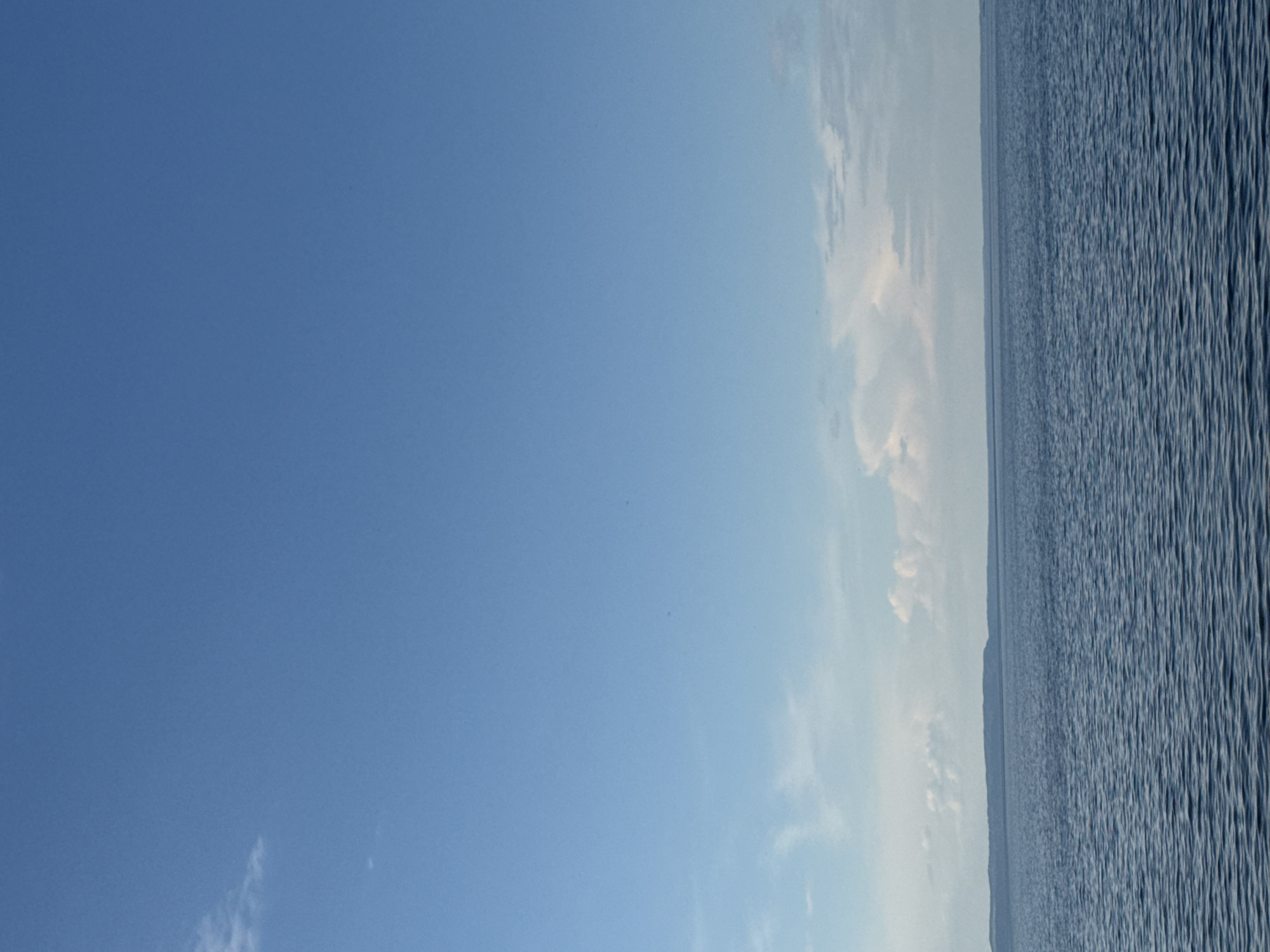A vertical, medium-distance, eye-level shot captures a serene, vast expanse of blue water meeting a distant, low-lying landmass under a clear blue sky with sparse clouds. The water exhibits a gentle ripple pattern, indicating a calm body of water, likely a lake or river. The landmass on the horizon is a muted blue-green, suggesting vegetation and a gentle, undulating terrain. A few wispy white clouds are scattered across the upper portion of the sky. The scene is devoid of any human presence or man-made objects, creating a natural and tranquil atmosphere. The image is consistent with a view of the Kama River Reservoir near Chaikovsky, Russia, during daylight hours with fair weather.