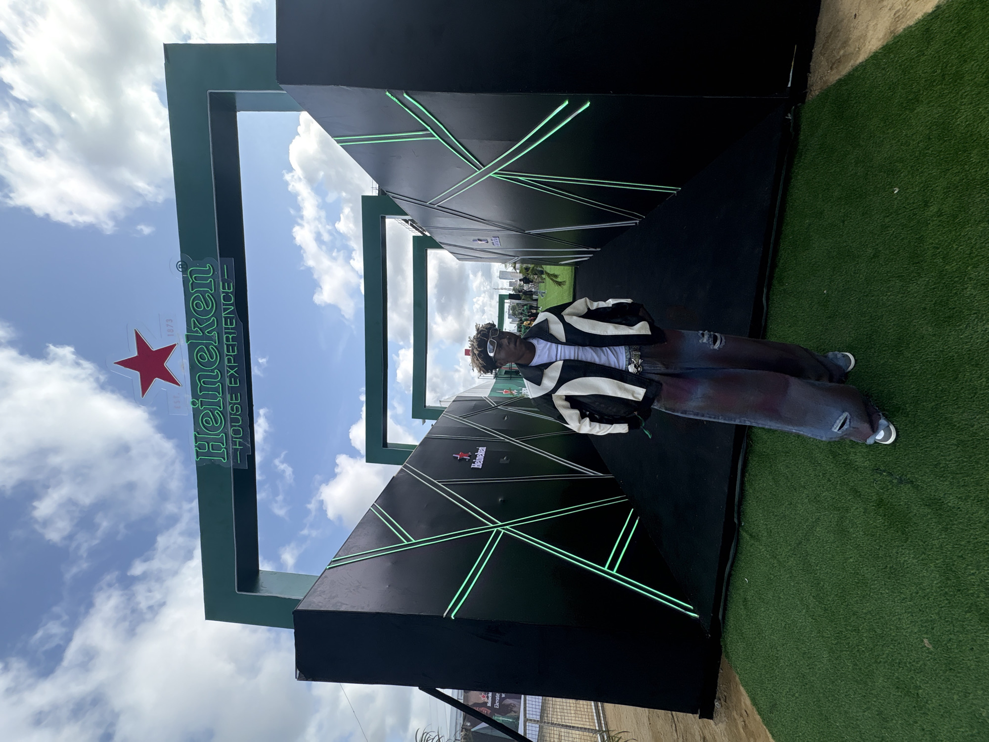 A man stands in front of a Heineken House Experience display in Lagos, Nigeria. The display features a large green Heineken logo with a red star and the words "House Experience" in white text. Behind him, the display is constructed from black panels with intersecting green neon lights. He is wearing a black and white jacket, a white t-shirt, and dark jeans. He has sunglasses on. The environment includes a blue sky with clouds and artificial green grass surrounding the display.