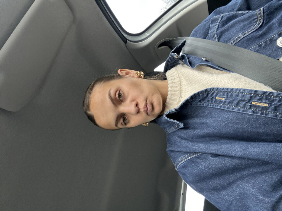 A light-skinned adult female is depicted seated within a vehicle, identified as being in Saint-Cyr-sur-le-Rhône, France. She is positioned to face the viewer directly, exhibiting a slight pout with her lips. Her light brown hair is pulled back from her face. She wears a cream-colored knitted sweater layered under a dark blue denim shirt or jacket featuring visible light-colored stitching and buttons. Ornate, gold-toned stud earrings with a textured design are present on both earlobes. A black seatbelt is securely fastened across her chest and right shoulder. The vehicle's interior consists of a light grey ceiling and a sun visor partially visible above her. A car window on the right side of the frame reveals an overcast sky and indistinct outdoor foliage, possibly bare trees or bushes.