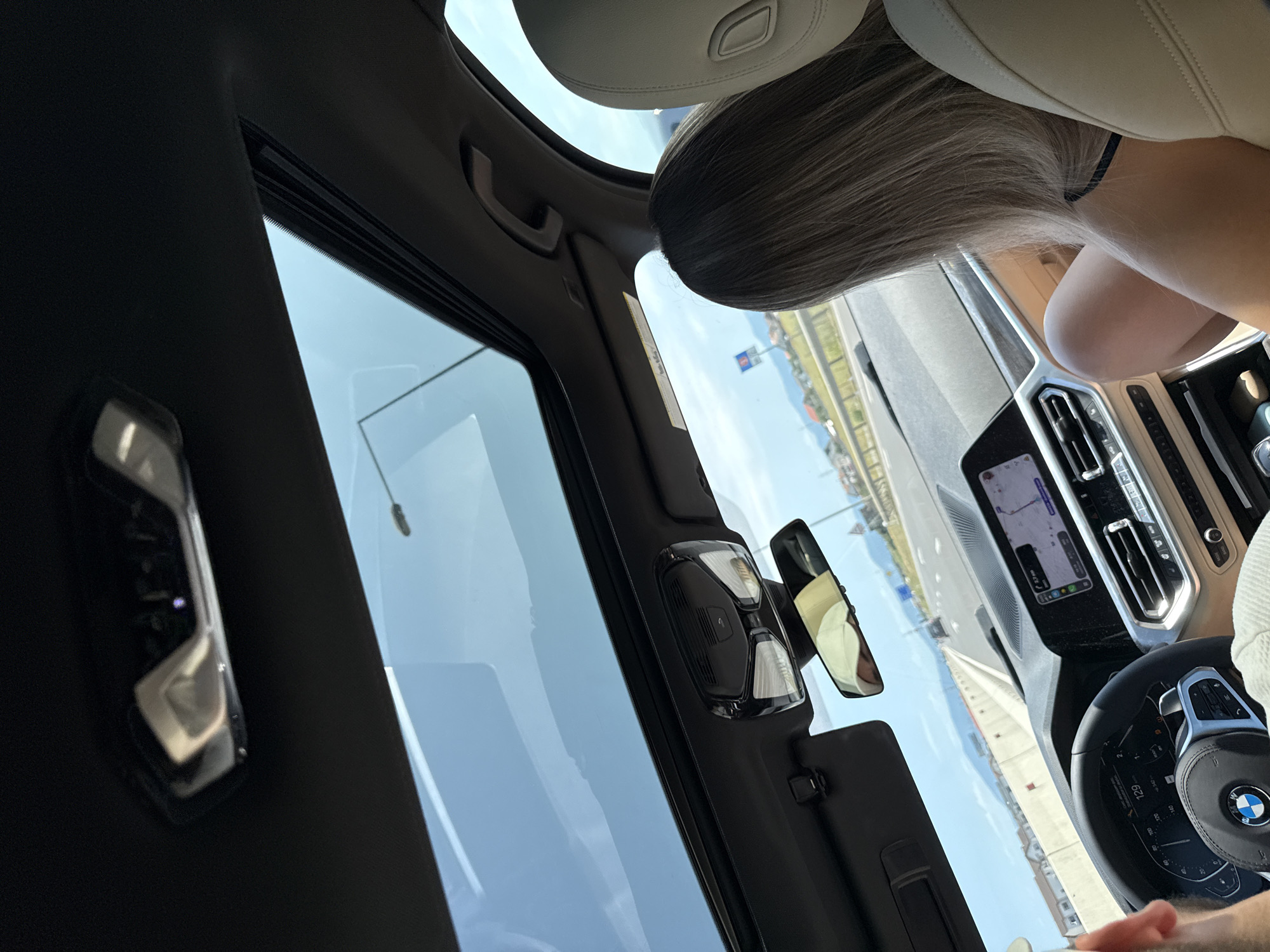 The image is a first-person view from inside a modern car, looking towards the dashboard and out the sunroof. The car is in motion on a highway, with a clear sky and distant mountains visible. The interior of the car features a panoramic sunroof, a digital dashboard displaying 129 km/h, a central infotainment screen with navigation, and tan leather seats. A person with blonde hair is seated in the passenger seat, facing forward. Another person's arm is visible on the left side of the frame, likely the driver. The scene appears to be on a highway near Sanpetru, Romania, as indicated by the environment and potential road signage, though no specific landmarks are definitively identifiable within the frame.