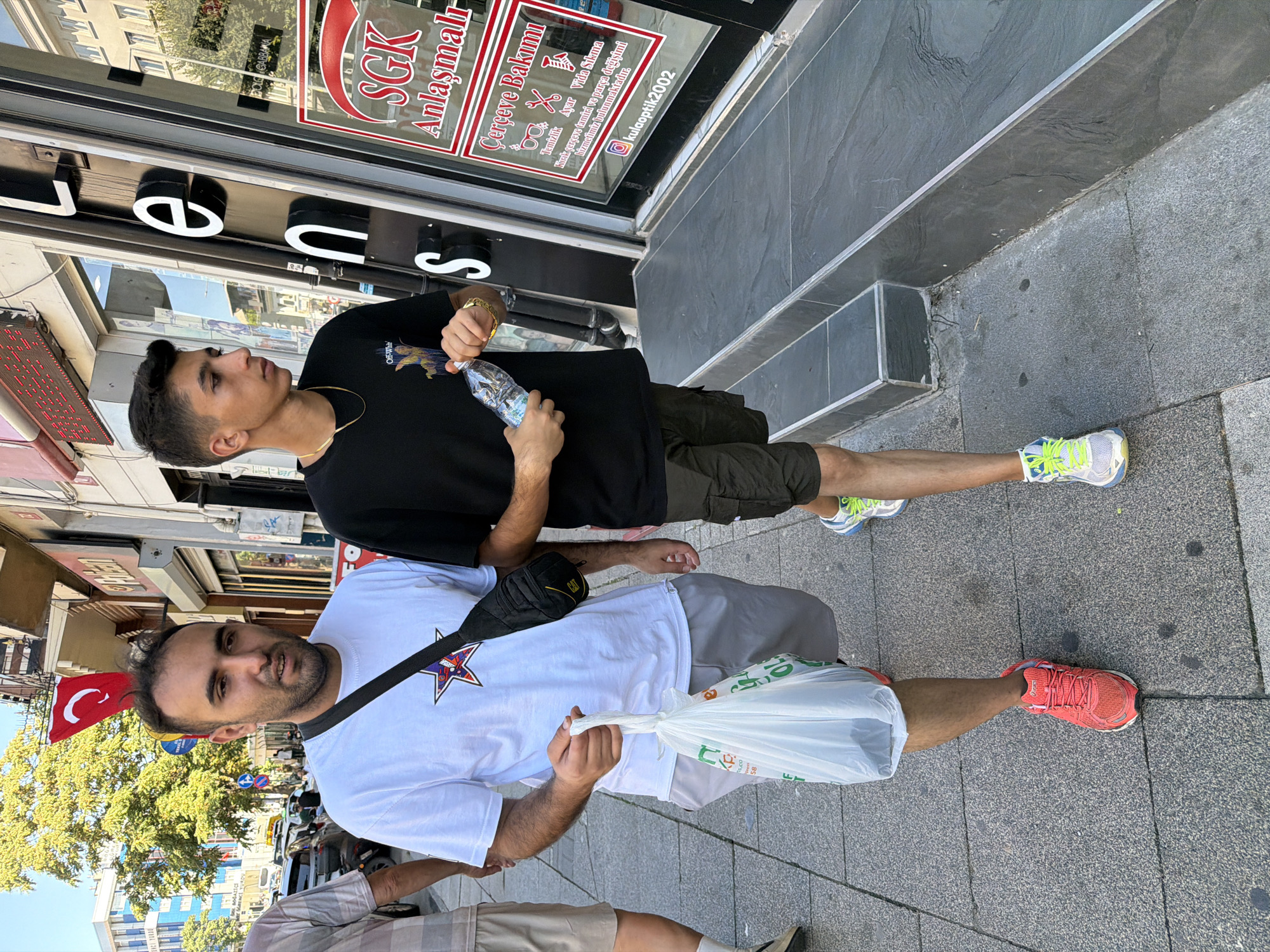 Two men are walking on a sidewalk in Kadıköy, Turkey. The man on the left is wearing a white t-shirt and gray shorts, carrying a white plastic bag in his right hand. He has a black fanny pack slung across his chest. The man on the right is wearing a black t-shirt and dark shorts, holding a bottle of water. They are walking past a store with a large sign that reads "LENSS" and a window displaying optical products and services. The street is lined with buildings and trees, and there are cars parked along the side of the road. A Turkish flag is visible on a pole in the background. The sidewalk is made of stone tiles, and there are steps leading up to the store entrance.