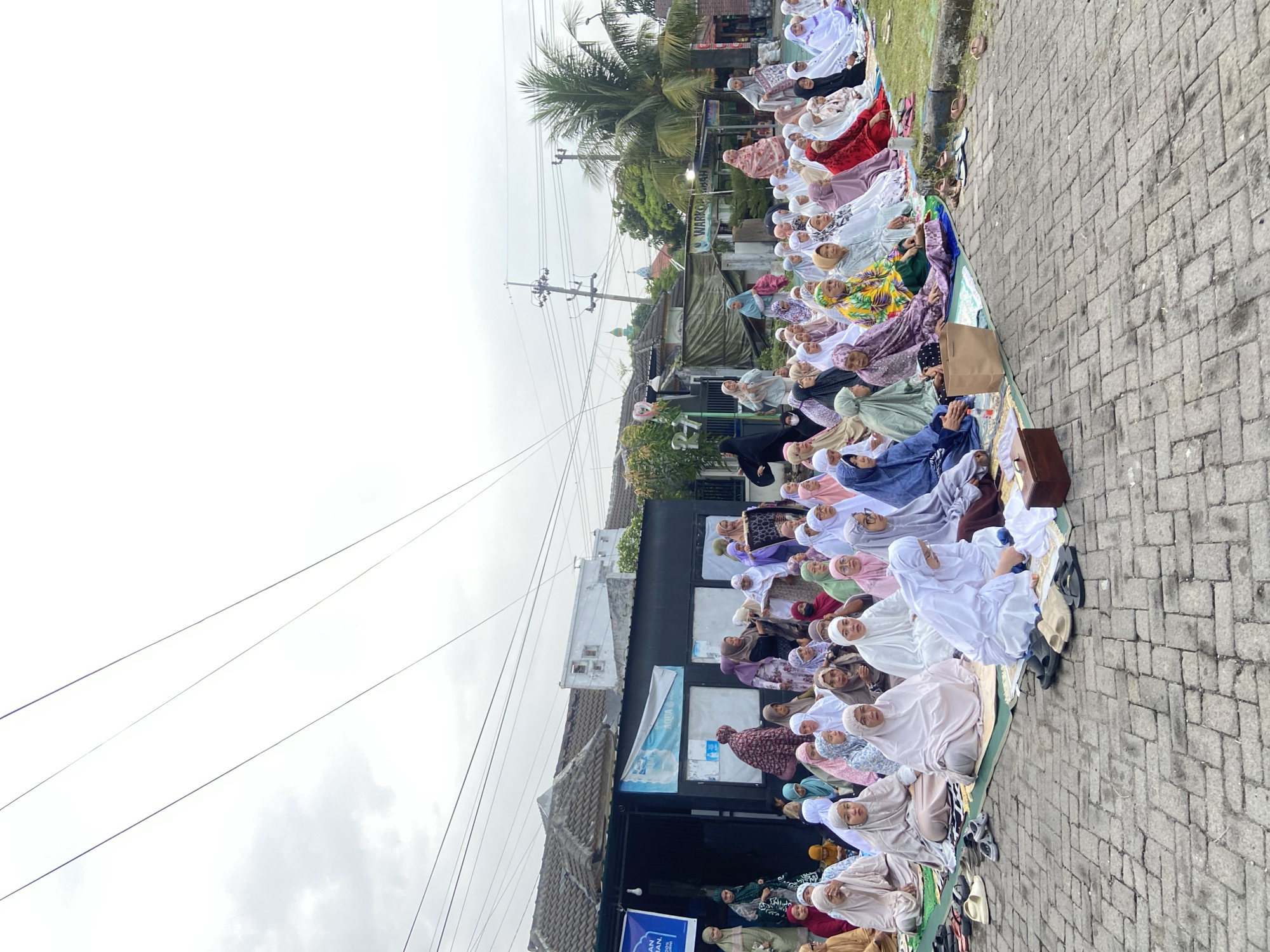 A medium shot, taken from the ground, shows a large group of women praying in a row outdoors, apparently on a city street in Gelam, Indonesia. The women are seated on mats or prayer rugs, wearing various colorful patterned Muslim headscarves and robes. They face to the right of the shot, toward a small building with advertisements posted on it. Overhead are power lines. The sky is gray and overcast. The ground is paved with bricks. In the background are trees.