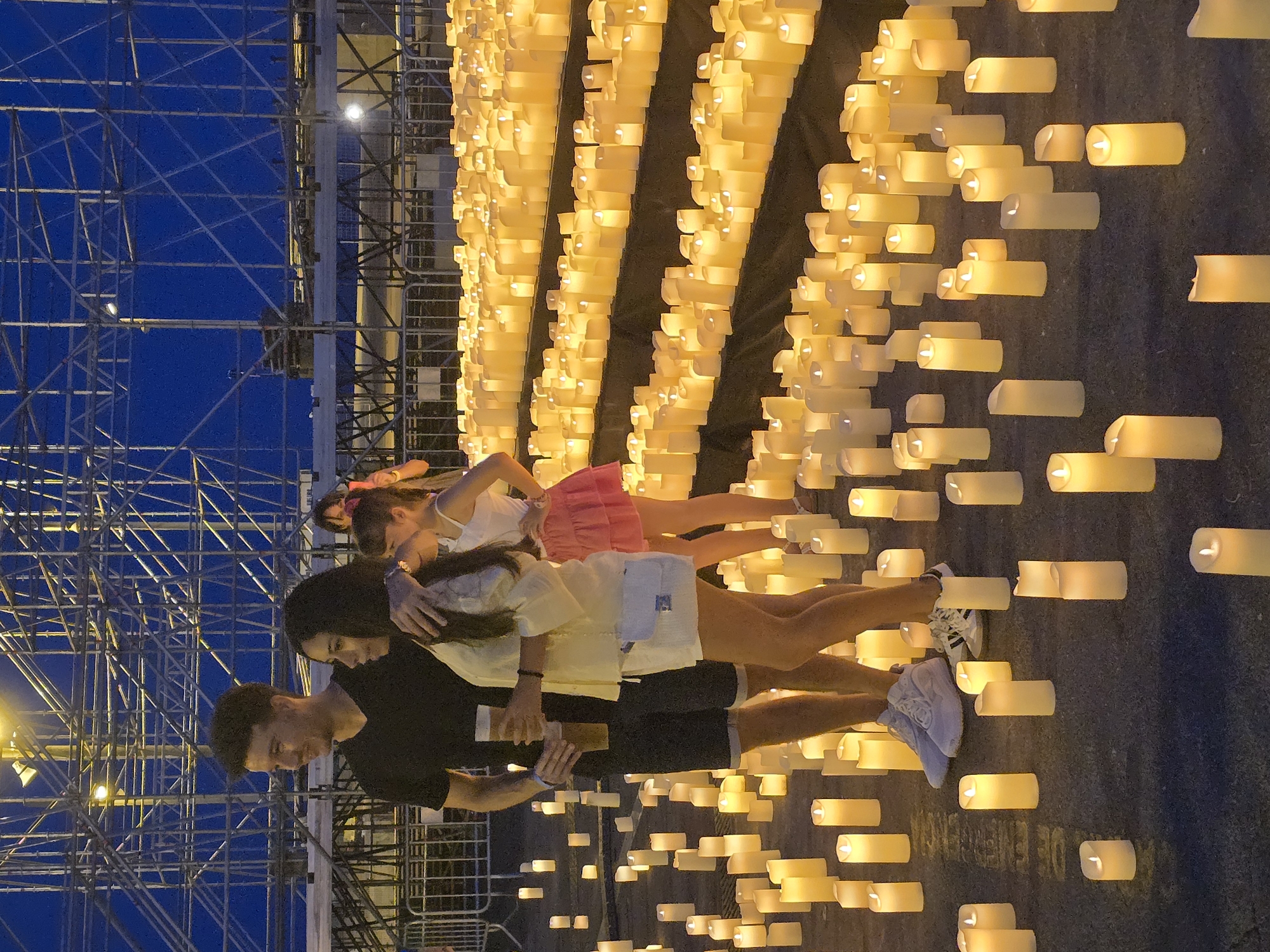 In Sant Adrià de Besòs, Spain, a man and woman stand amidst an expanse of lit candles at dusk. The couple is positioned in the foreground, with the man on the left embracing the woman. He wears a black t-shirt and dark shorts, holding a beer in his right hand. The woman, on the right, is in a white top and shorts, with a small white crossbody bag. Behind them, a young girl in a white top and pink skirt stands. The ground is covered with countless white LED candles arranged in rows, creating a luminous carpet that extends into the background. In the distance, scaffolding and a dark sky suggest an urban or event setting. The lighting is dim, with the primary illumination coming from the candles.