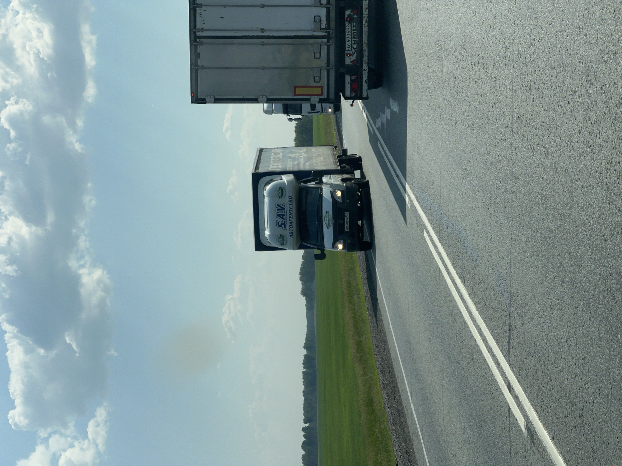 The image depicts a view from a vehicle traveling on a paved road in Chik, Russia, during daytime. The sky is a clear blue with scattered white clouds, suggesting fair weather. The road has visible white lane markings.
The primary subjects are two large trucks. In the foreground, a white truck with a black cab is visible. The side of its trailer displays text, including "S.A.V." and "АВТОАГЕНТСТВО". A license plate is visible on the front of the cab, reading "H930PY 102". Behind this truck, a larger, silver-colored truck with a white trailer is seen. The rear of this truck has visible license plates, one reading "Aa 9705 42", and "SCHMITZ" branding. A smaller white truck is visible between the two larger trucks.
The scene is set along a highway with a strip of green grass and a line of trees visible on the left side of the road, indicating a rural or semi-rural landscape. The orientation of the image suggests it was taken from a car traveling behind or alongside the trucks. There are no people clearly visible in the image. The overall impression is one of a journey on a highway.