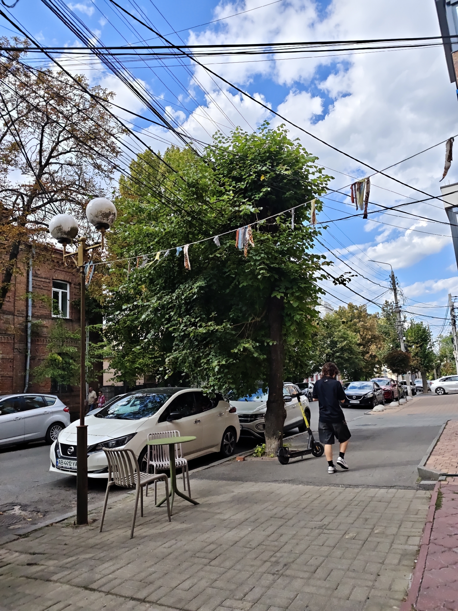 This is an eye-level, medium shot of a tree-lined street in Vinnytsia, Ukraine, on a cloudy day. The foreground features a brick sidewalk with a modern green table and two chairs. A white electric car is parked on the street next to a light pole with two spherical lamps. Further down the street, several other cars are parked, and a person in a black t-shirt and shorts is walking away from the camera, with an electric scooter on the ground beside them. Buildings and trees line the street, with power lines crisscrossing overhead. Decorative fabric strips are hung from some of the power lines.