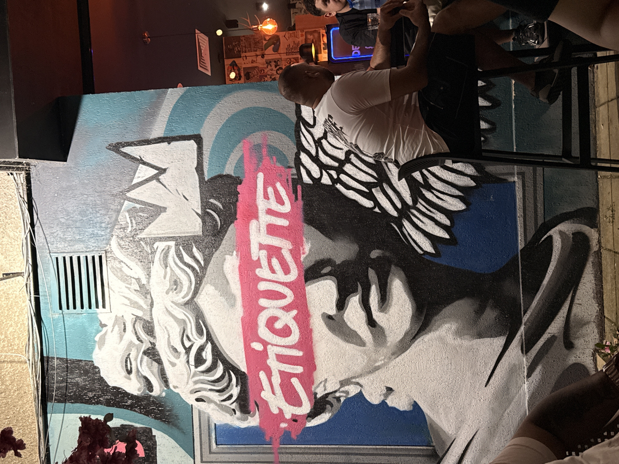 The image depicts a mural on a wall, featuring a black and white bust of a classical figure with a pink brushstroke across its eyes, on which the word "ETIQUETTE" is spray-painted in white. Above the figure's head is a stylized crown. The mural is located on an exterior wall, with electrical conduits and a ventilation grate visible above it. To the right of the mural, a portion of an indoor space is visible, with people sitting at tables and hanging light fixtures. One man, wearing a white t-shirt and dark shorts, is seated on a high stool in the foreground, facing away from the camera. Another man, wearing a black shirt, is partially visible to his right. The setting appears to be a bar or restaurant. While the image is taken in Lovech, Bulgaria, specific details about the location within the city are not evident from the visual information provided.