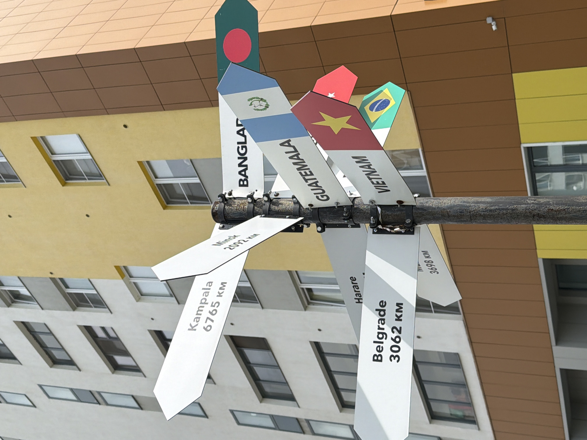 A directional signpost with multiple arms points to various international cities from a city location in Yekaterinburg, Russia. The signpost has white arms with black text indicating city names and distances in kilometers, and some arms are decorated with national flags. Some of the visible destinations include Kampala, Minsk, Harare, Belgrade, Bangladesh, Guatemala, and Vietnam. The signpost is mounted on a dark, weathered pole. In the background, a modern building with a beige and yellow facade, featuring multiple windows, is visible. The scene appears to be outdoors during daylight.