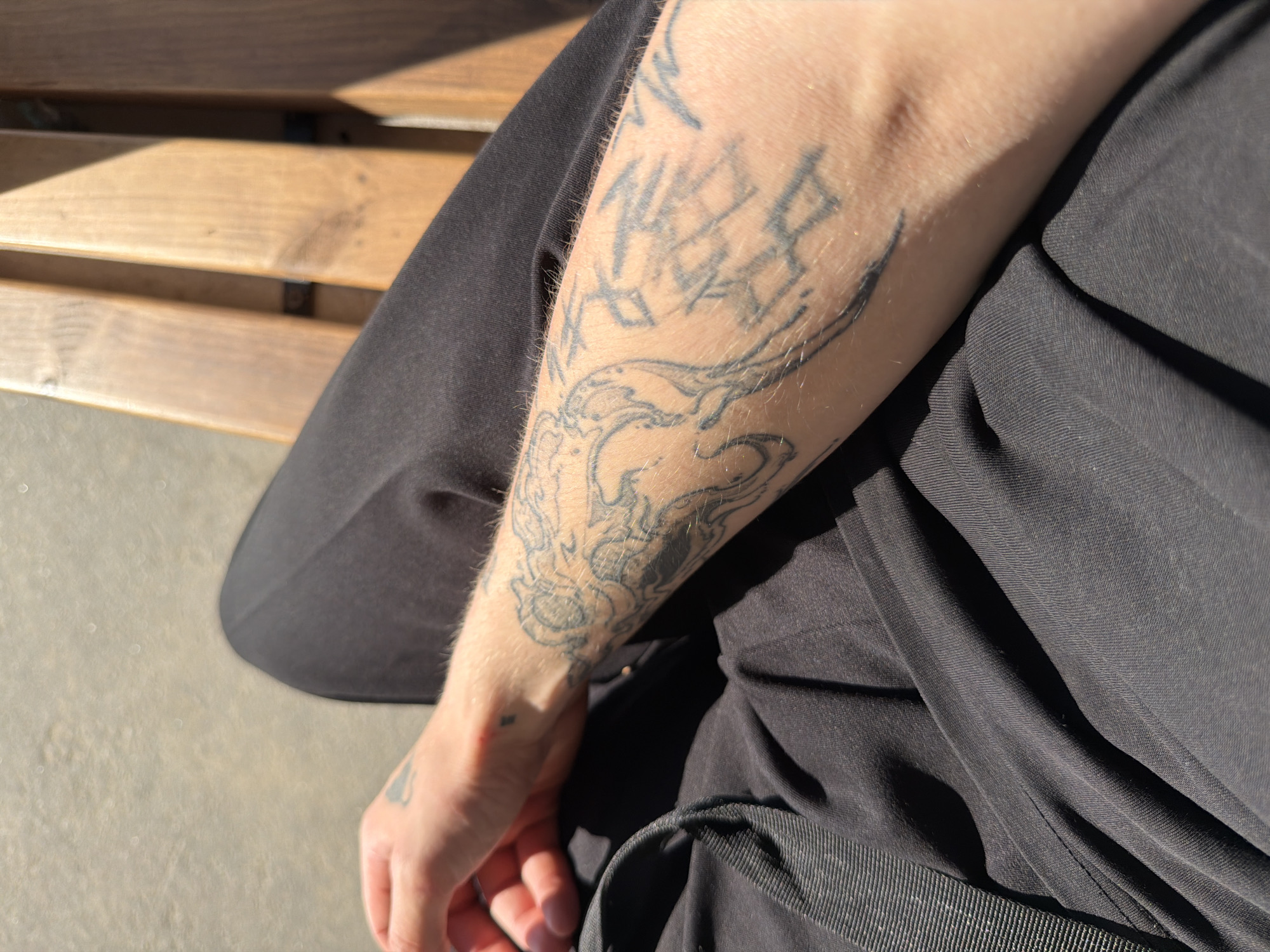 In Priovrazhnoe, Russia, a person sits on a wooden bench outdoors. Their left arm, adorned with tattoos, rests on their lap. The individual is wearing dark clothing, and the scene is illuminated by bright sunlight, casting shadows on the bench. The ground appears to be a light-colored gravel or dirt path.