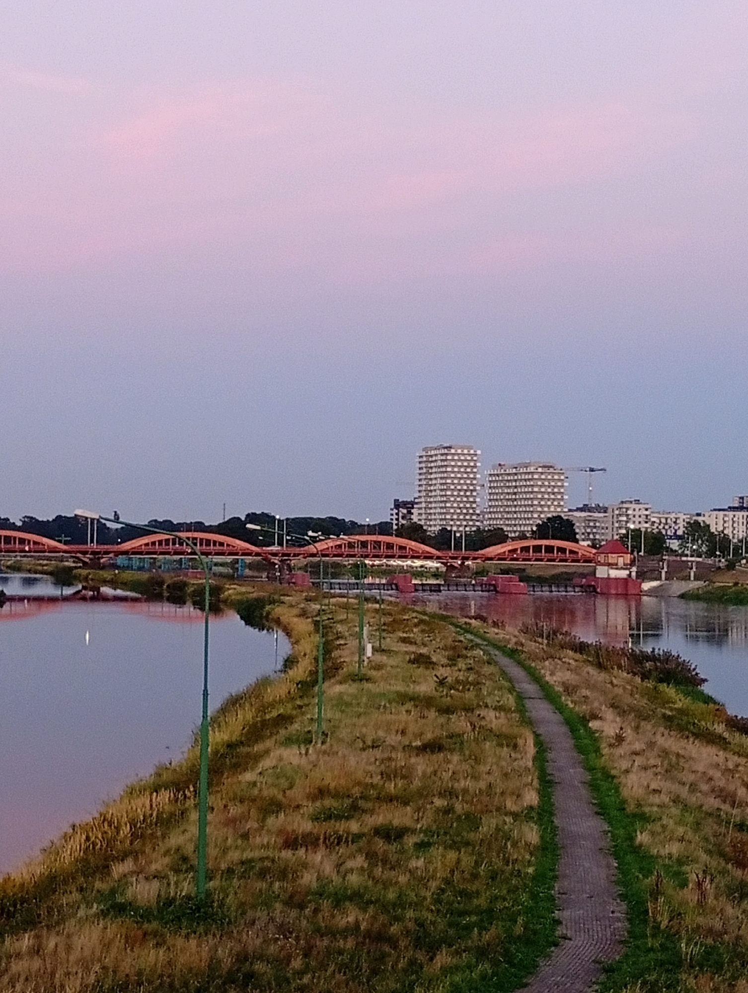 The image shows a view of the Oder River in Wrocław, Poland, at dusk. The sky is a soft pink and purple, indicative of sunset or sunrise. A curved, orange bridge spans the river in the middle ground. On the far bank, a city skyline is visible with several modern, high-rise buildings. In the foreground, a grassy embankment slopes down to the water, with a paved pathway winding along the top. Several green lampposts are scattered along the embankment. The water of the river is calm and reflects the colors of the sky and the bridge. The scene appears peaceful and serene.