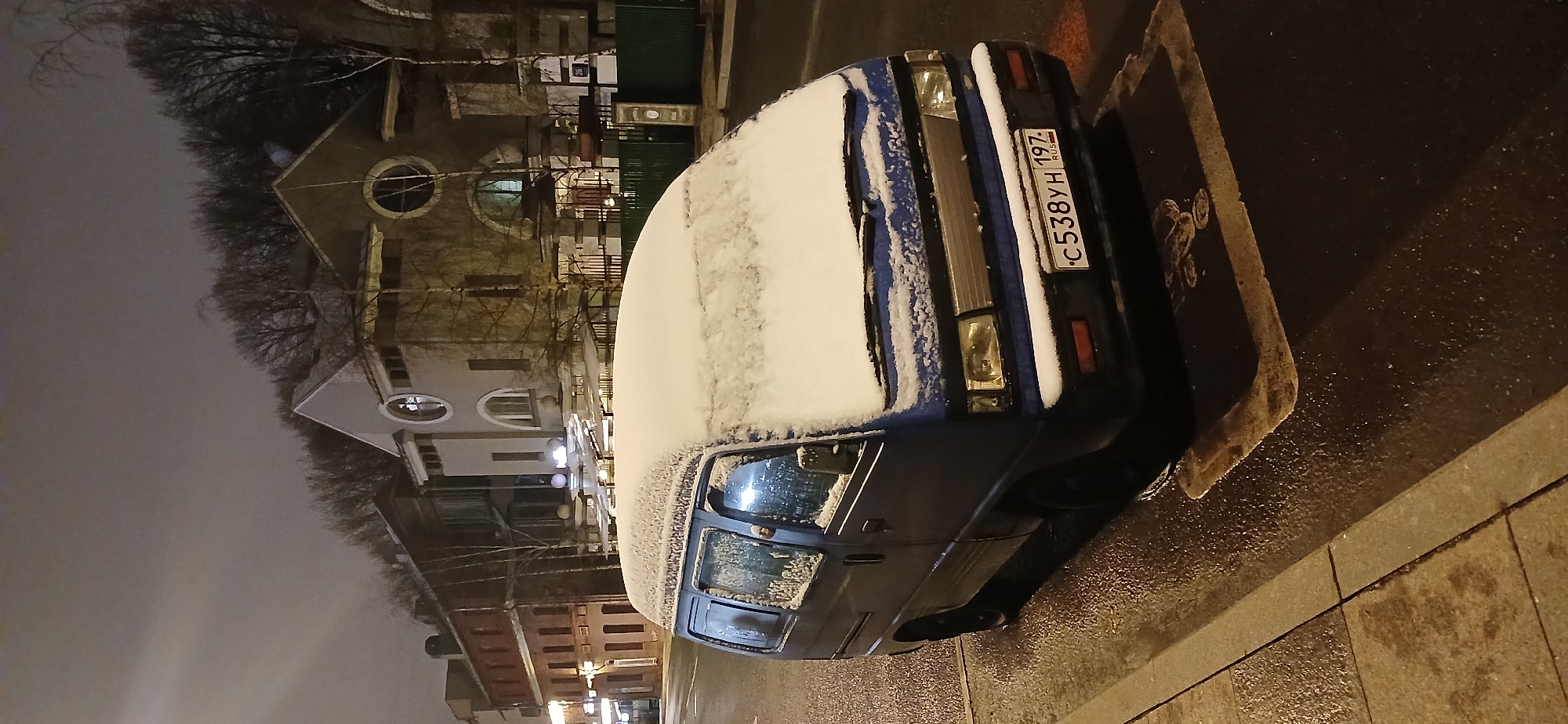 A blue minivan, partially covered in snow, is parked on a wet street next to a sidewalk. The vehicle has a Russian license plate that reads "C538YH 197 RUS." The background features a building with a distinctive circular window design. The photograph was taken at night, likely in Moscow, Russia, due to the vehicle registration code "197," which is associated with Moscow. There are no people visible in the image.