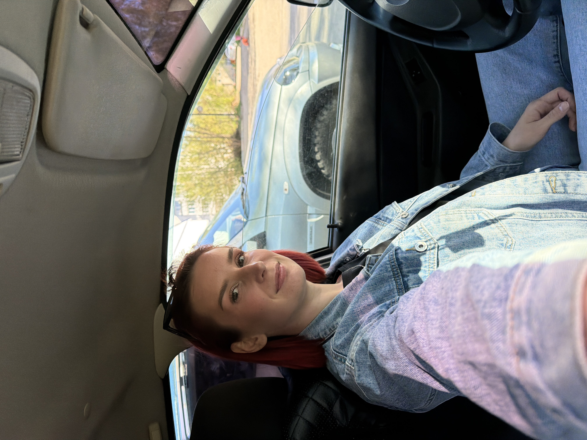 A young woman with red hair, wearing a denim jacket and jeans, is sitting in the driver's seat of a car. She is smiling and looking at the camera. Through the car window, another car and a building are visible. Based on the buildings and the car visible, this may be Chaikovsky, Russia.