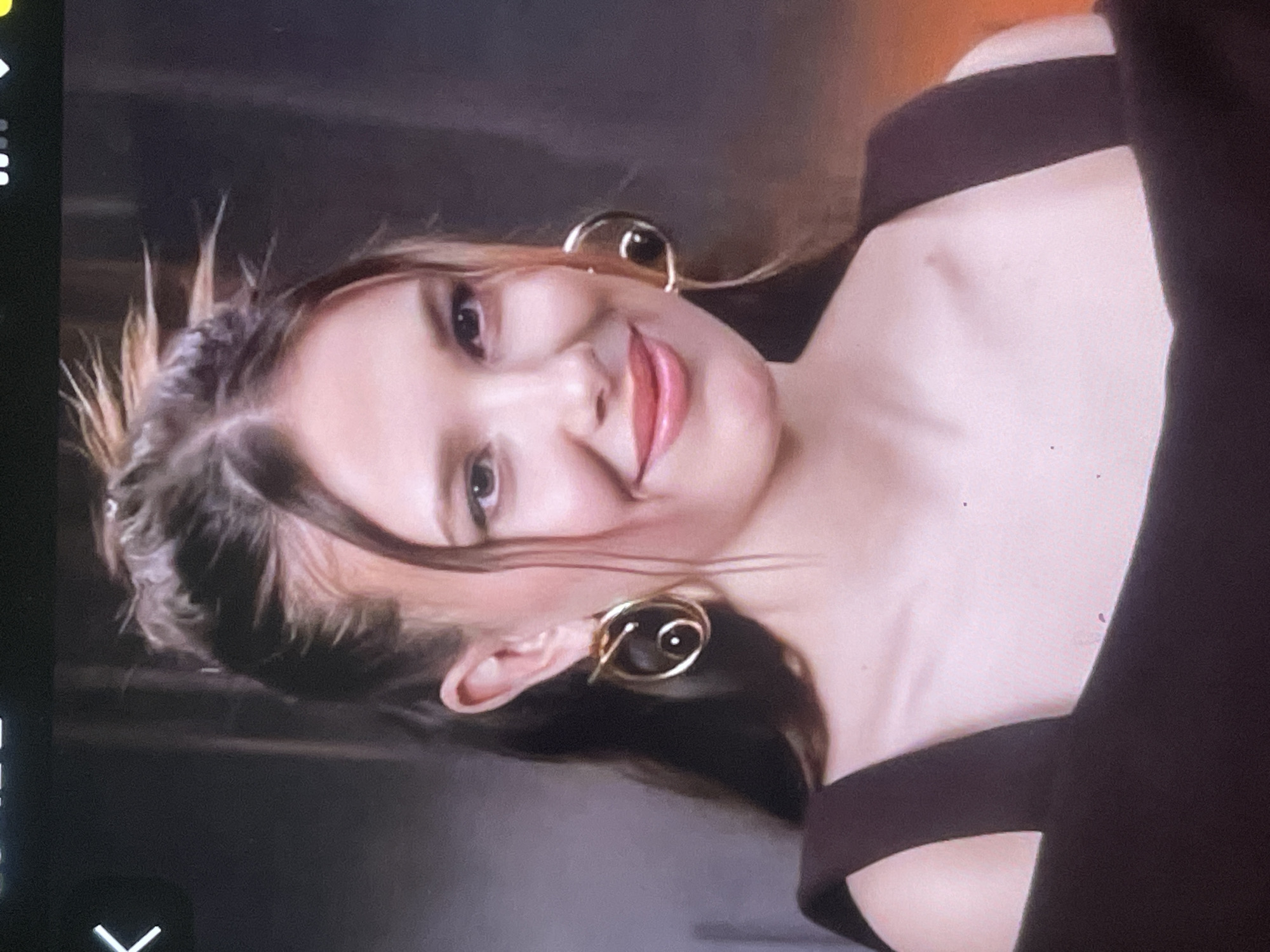 A young woman, likely in her late teens or early twenties, is depicted in a close-up portrait, smiling gently at the camera. She has fair skin, dark eyes, and light brown or blonde hair styled in a messy updo with wispy strands framing her face. Her lips are adorned with light pink lipstick.
She is wearing prominent, large, gold-toned spiral or swirl-shaped earrings on both ears. Her attire consists of a dark-colored top, possibly black or dark brown, with a broad strap visible over her shoulder and a cut-out or multi-strap design across her chest.
The background is dark and out of focus, suggesting an indoor setting with atmospheric lighting. A subtle warm glow is present in the upper right of the frame, possibly from an indirect light source. The woman's head is slightly tilted, conveying a relaxed and approachable demeanor.
On the left side of the original image, digital interface elements (carets and dots) are visible, indicating the image might be a photograph of a screen or a screenshot. The overall impression is one of pleasantness and casual elegance. No specific visual cues in the image directly indicate the location of Poltava, Ukraine.