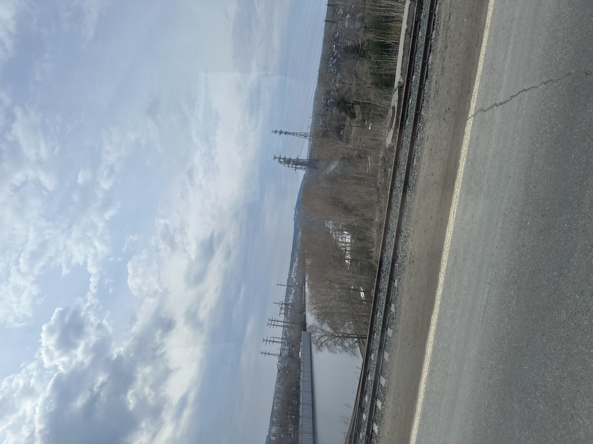 The image shows a view from an elevated position in the city of Chaikovsky, Russia, with a focus on the natural and industrial landscape. The sky is filled with clouds.
In the middle ground, there's a row of bare trees, power lines running across the scene, and distant buildings that are hard to discern. A body of water runs adjacent to the tracks.
In the foreground, railway tracks and an asphalt surface, likely a road or pathway, are visible.