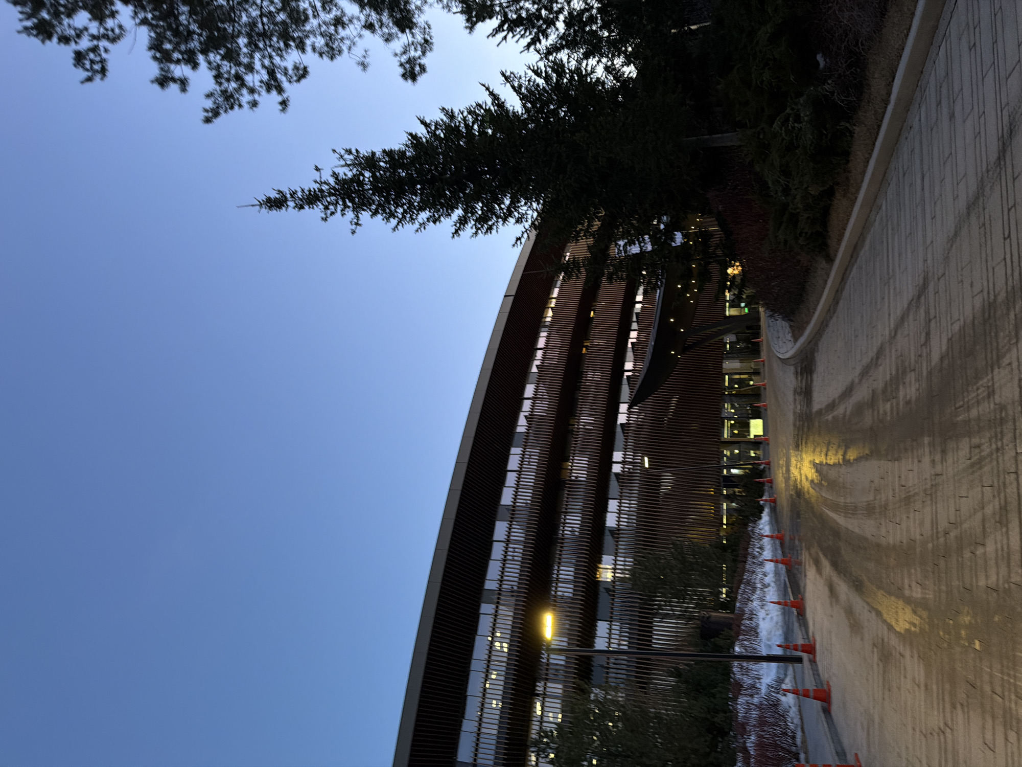 The image shows a curved modern building with horizontal wood-like slats against a blue sky. A tall evergreen tree is partially visible above the building. Along the side of the building is a paved walkway that appears wet, reflecting light. A line of orange cones is positioned between the walkway and a landscaped area. A lamppost illuminates a section of the building and the walkway. It is evening. The building's windows indicate lit interior spaces. The location is Озёрное, Russia.