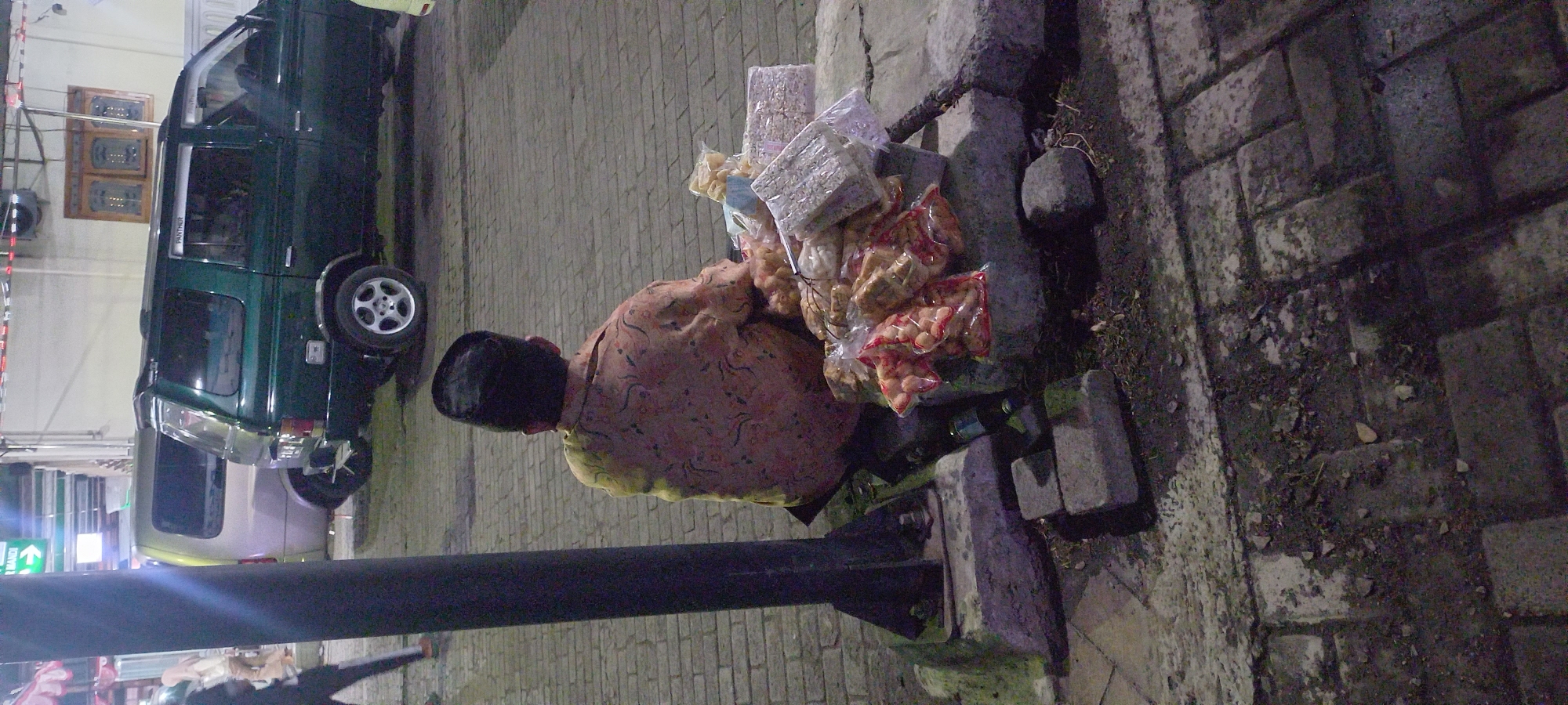 The image depicts a street scene in Banjarsari, Indonesia, at dusk or night. A person wearing a black peci cap and a patterned shirt is seated on the ground, facing away from the camera. They are positioned next to a pile of packaged snacks, which appear to be a variety of fried and extruded food items. The snacks are in clear plastic bags, some with red accents. The person is seated on a concrete block next to a dark, vertical pole. In the background, two vehicles are parked on a brick-paved surface: a green SUV and a lighter-colored van. The lighting suggests artificial illumination from nearby buildings or streetlights, casting shadows on the ground. The environment appears to be an urban street or sidewalk.