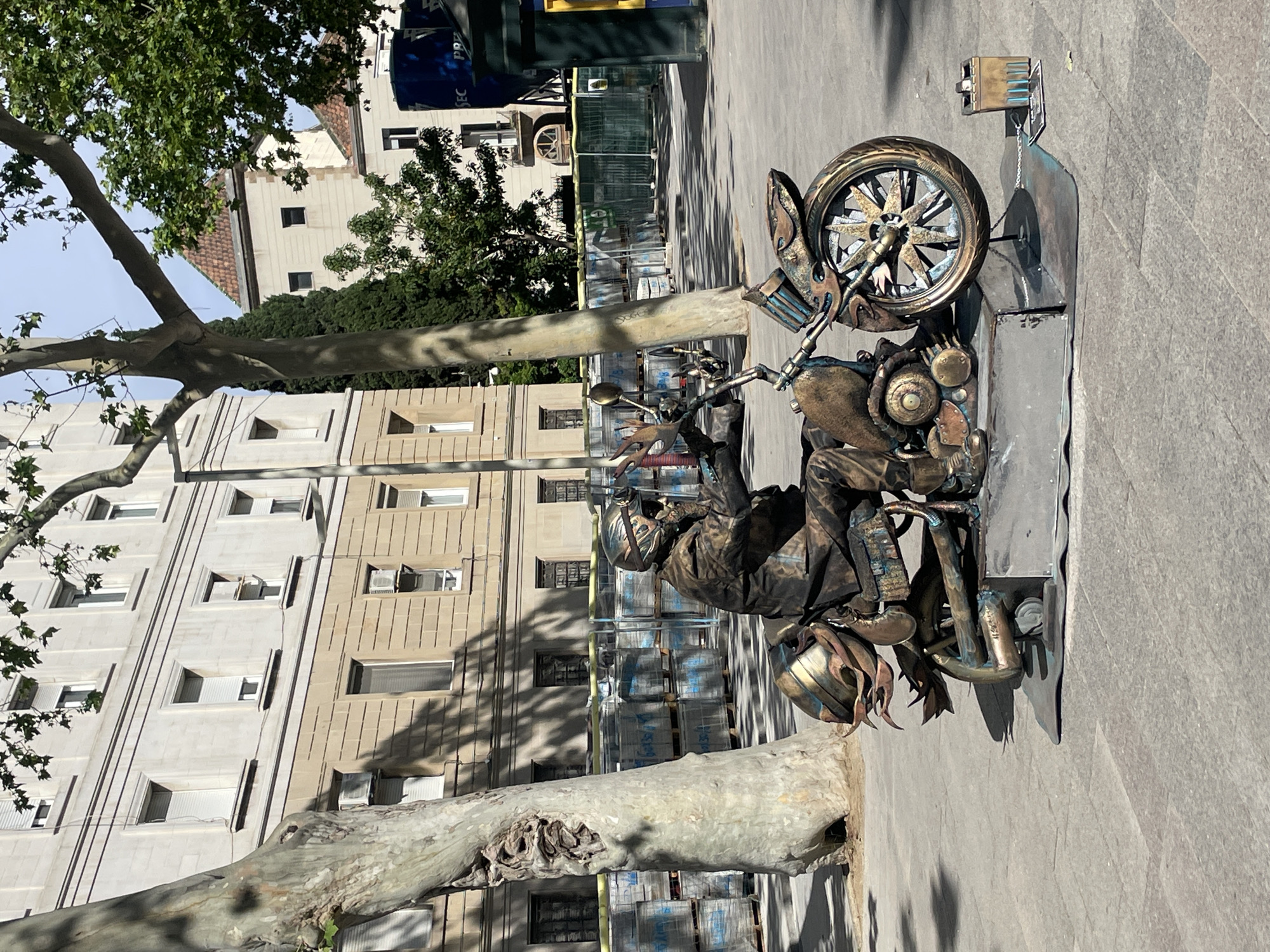 A vertical, full-shot photograph depicts a metallic sculpture of a person riding a motorcycle, set against a backdrop of buildings and trees in what appears to be a city square. The sculpture is prominently positioned on the right side of the image. The motorcycle has a detailed design, featuring intricate metalwork, with the front wheel prominently displayed. A metallic figure sits astride the motorcycle.
The environment includes a paved ground surface, indicative of a pedestrian area. Behind the sculpture, the architecture includes multi-story buildings. There are trees with visible branches, suggesting an urban planting within a square or park. The scene gives the impression of a public art installation in a city setting, likely in Barcelona, Spain.