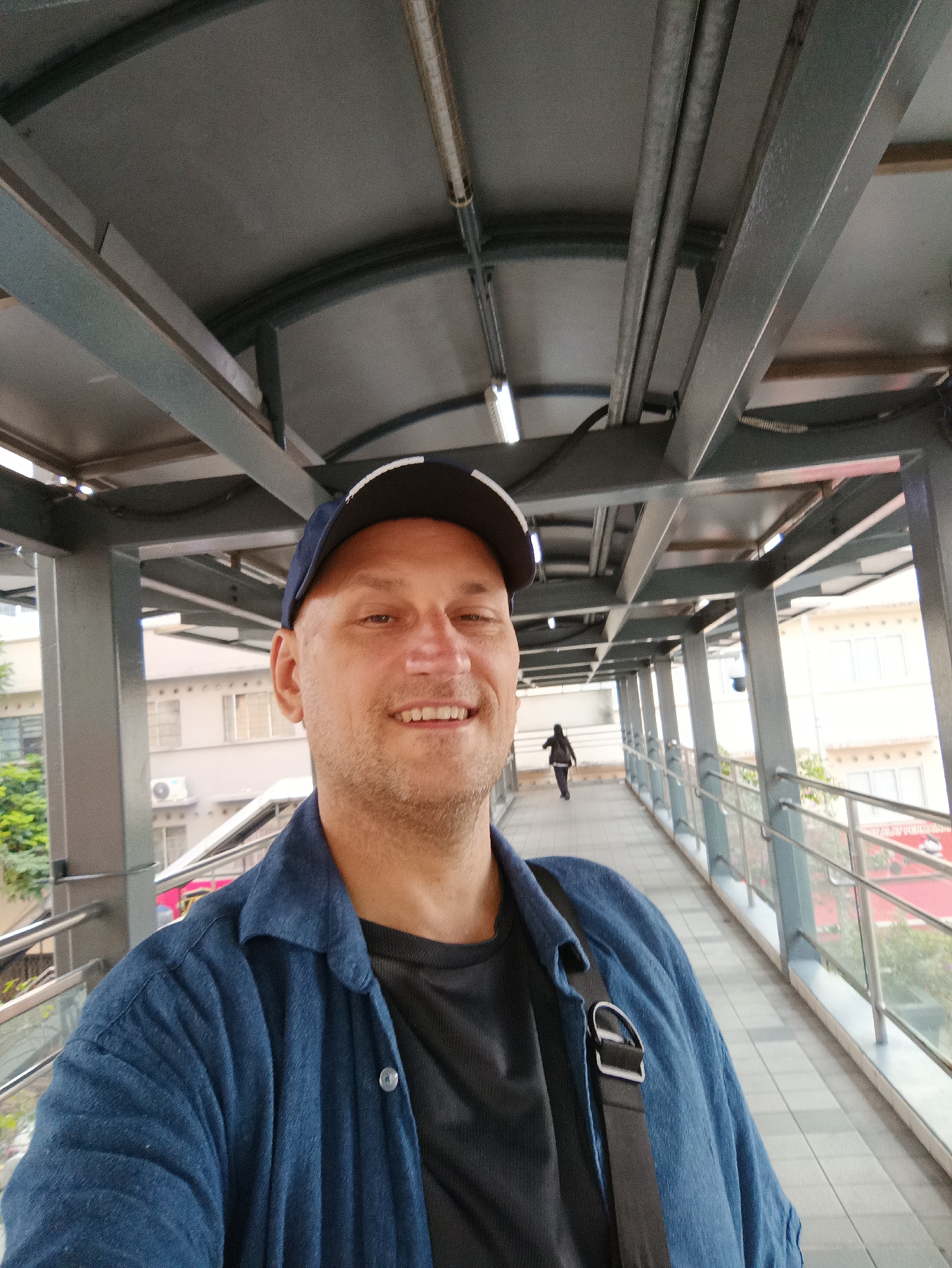 A man wearing a blue cap and a blue collared shirt over a black t-shirt poses for a selfie on a covered pedestrian walkway. The walkway has a metal structure overhead and railings along the sides, with buildings visible in the background. Another person is walking away in the distance on the walkway. The scene appears to be in an urban environment, possibly Kuala Lumpur, Malaysia, given the architectural style of the buildings and the presence of a covered walkway.