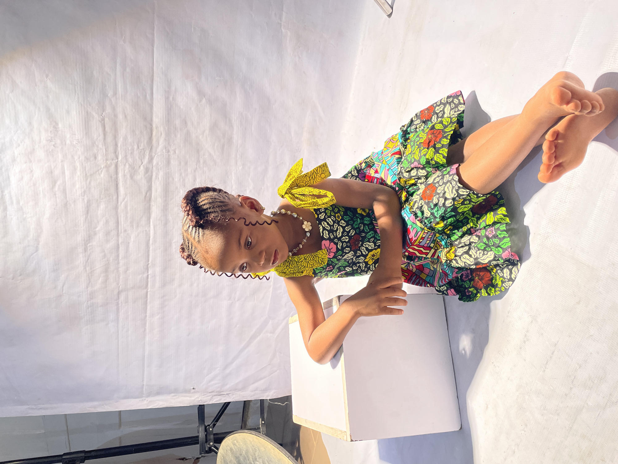 This portrait photograph, taken in Jos, Nigeria, features a young girl posing within a makeshift studio setup. The image depicts the girl seated on a white surface, with a plain white fabric backdrop behind her. She is leaning forward slightly, resting her left arm on a white rectangular box placed in front of her.
The girl has her hair intricately styled in braids, with several curly tendrils framing her face. She wears a vividly colored dress, rich with floral and abstract patterns in shades of green, yellow, red, and blue. The dress is characterized by yellow bow-like embellishments on its shoulders. Around her neck, she wears a white beaded necklace with a small flower charm. Her bare feet are visible, crossed at the ankles, resting on the white surface. She looks directly at the camera with a focused and serene expression.
The scene is bathed in strong, natural sunlight, casting distinct shadows from the girl and the white box, indicative of a bright, sunny day, likely around midday. The white backdrop, which also covers the floor, appears to be a creased fabric sheet. In the lower left corner of the frame, parts of black metallic support structures, possibly from a stand holding the backdrop, are visible, along with a partial view of a drum or a similar round object. The overall impression is a charming and carefully composed, yet informal, photoshoot.