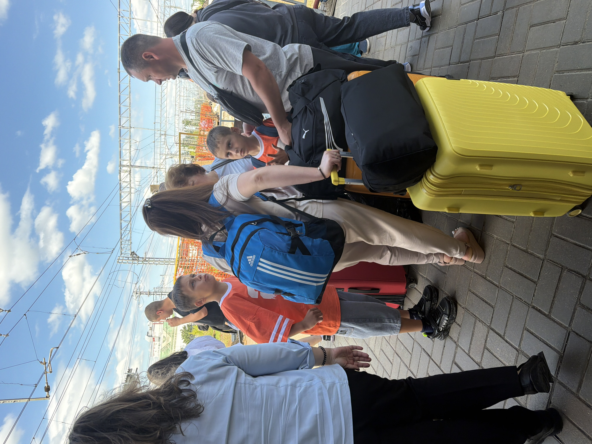 At a train station, a group of people are waiting on a paved platform under a blue sky with scattered clouds. A woman with long dark hair in a light blue shirt and black pants stands on the left, her back to the camera. Beside her, a boy in an orange shirt and denim shorts looks up and to the right, holding his right hand up. He is standing next to a red suitcase. A woman in beige pants and a white shirt, wearing a blue backpack with white stripes, is to his right. A man in a grey shirt and dark pants stands to the right of the woman in beige, holding a black bag with a white logo. Another boy in an orange shirt and denim shorts is partially visible next to the man. In the foreground, a bright yellow suitcase with a black duffel bag on top is positioned on the ground. Overhead, electrical wires and structures of the train station are visible against the sky.