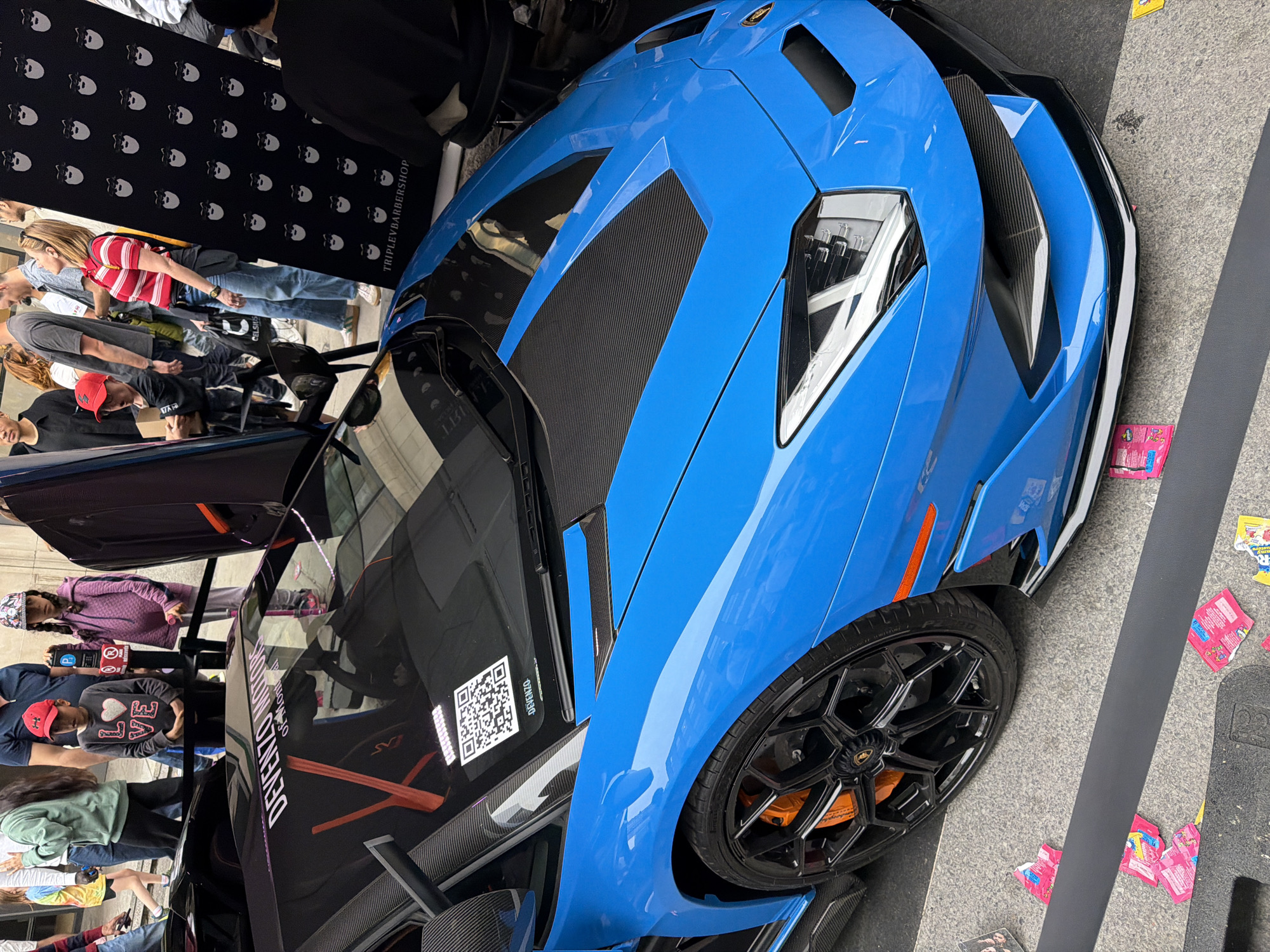 The image depicts a bright blue Lamborghini sports car parked on a street. The car has black rims, orange brake calipers, and carbon fiber accents on the hood and side. A QR code sticker is visible on the car's side window, along with the text "DEVENZO MOTOR of Toronto". The car's door is partially open, revealing an orange interior.
Several people are visible in the background, some looking at the car, suggesting the car is on display or part of an event. A black banner with skull motifs is also visible in the background, possibly indicating a brand or sponsor. The street surface appears to be concrete or asphalt. The presence of numerous candy wrappers littering the ground could indicate a playful event or gathering.
Although the specific location cannot be identified from the image, the text on the window suggests it's likely an event involving Devenzo Motor of Toronto, showcasing the blue Lamborghini. Given there is nothing to specifically identify this, the location in Montreal is not possible to confirm.
