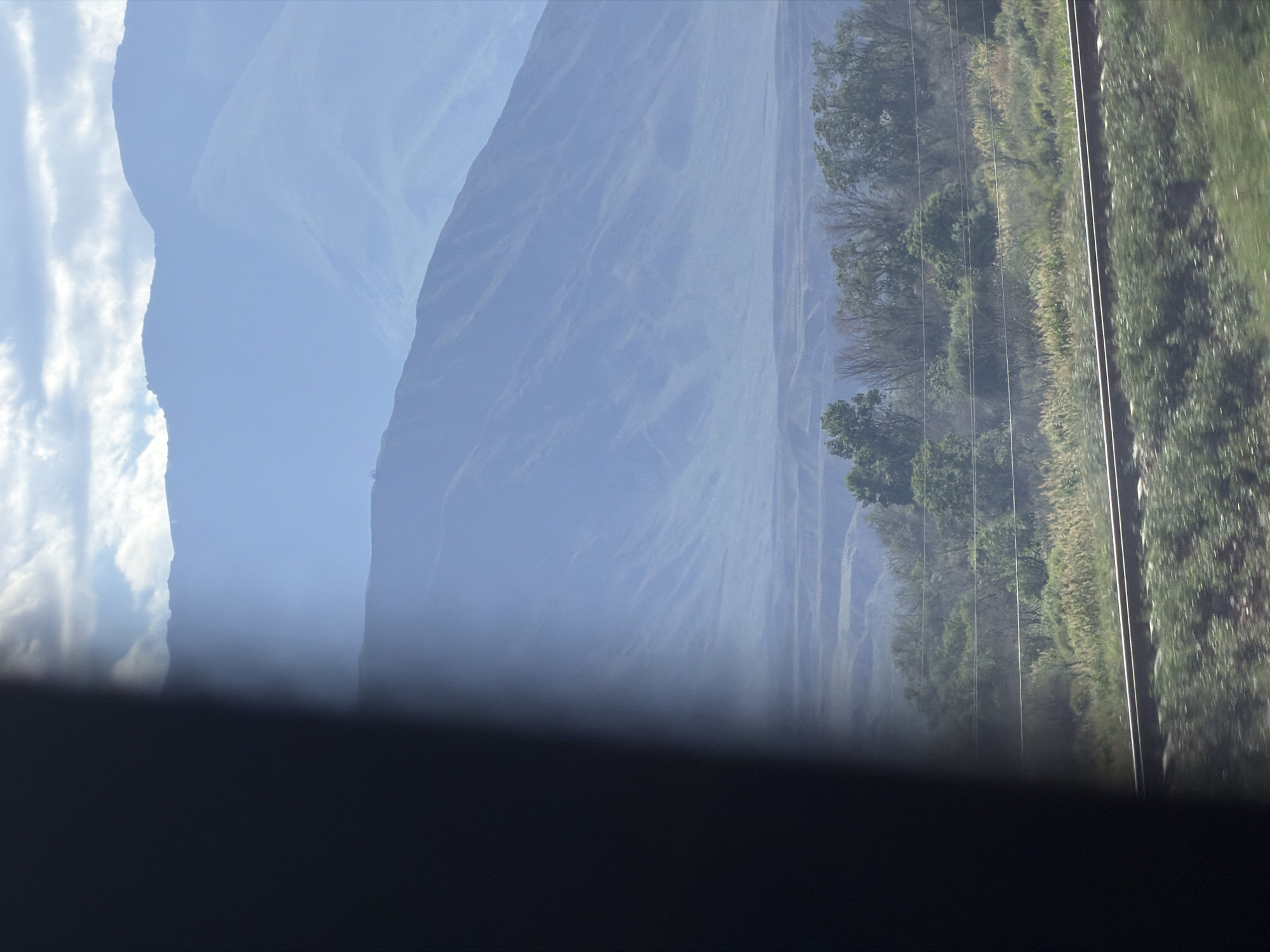 The image captures a distant mountain range veiled in a soft, blue haze. The mountains recede into layers, with the closest range showing more distinct contours and vegetation, while the further mountains are mere silhouettes against a pale sky. In the foreground, a blurry section of dark, vertical material, possibly a window frame or a structural element, partially obscures the view on the left. To the right of this dark section, a landscape unfolds. Tracks of a railway line run horizontally across the lower portion of the image, with blurred green foliage in front of them. Beyond the tracks, a band of trees and shrubbery creates a textured middle ground. The overall scene evokes a sense of travel and observation, likely from a moving vehicle. The location of Kok Moynok Eki, Kyrgyzstan, is a rural or mountainous area, consistent with the depicted scenery of rugged terrain and open skies.