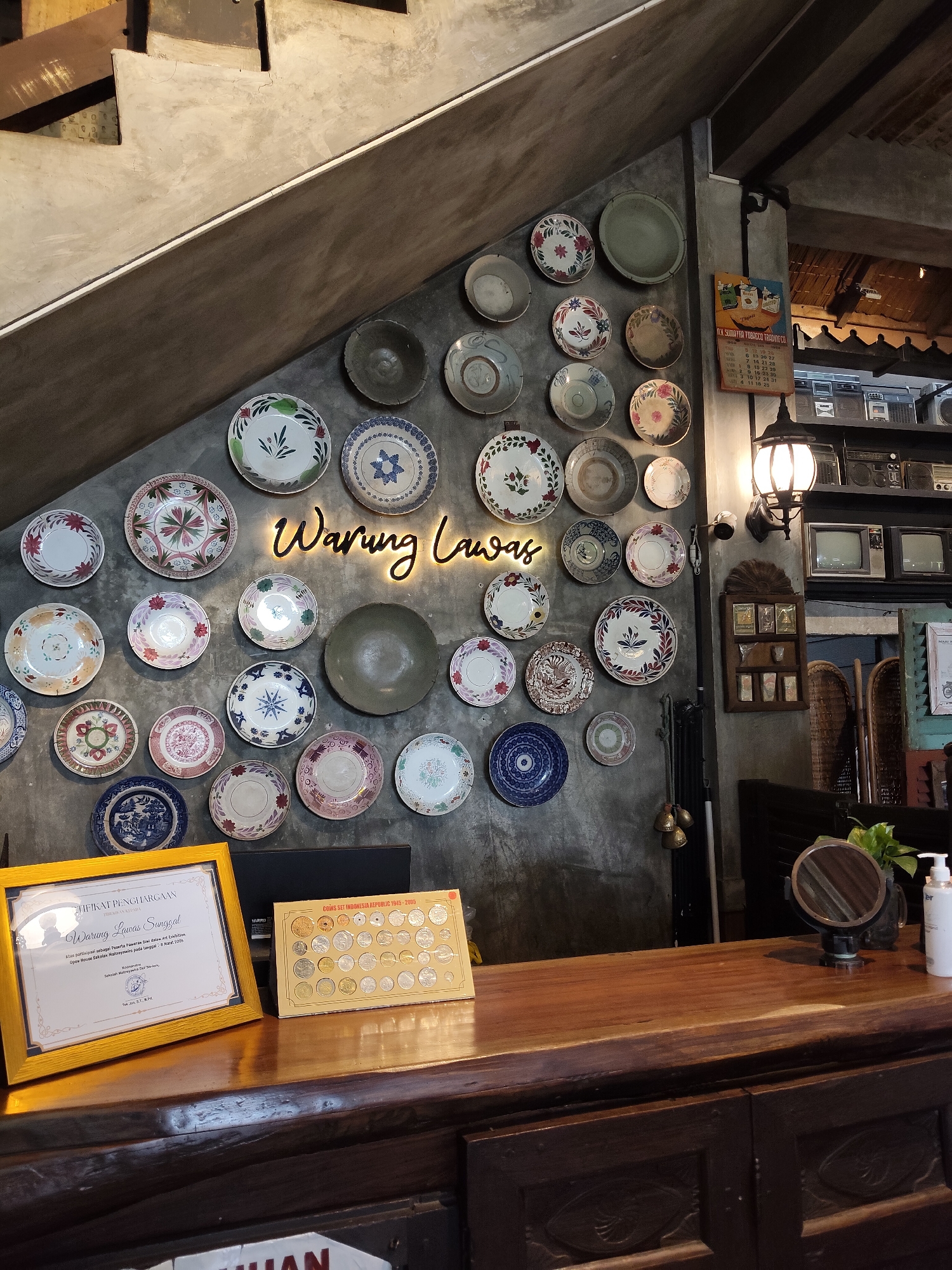 The image depicts the interior of Warung Lawas, a restaurant located in the City of Medan, Indonesia. The scene is characterized by a rustic and vintage aesthetic. The wall behind a wooden counter is adorned with an array of decorative plates, some with floral patterns and others with intricate designs. A neon sign spelling "Warung Lawas" in a script font glows on the wall. To the right of the plates, a shelf displays vintage electronics, including several boomboxes and televisions, as well as a framed calendar. A black lantern hangs from the wall, casting light on the area.
On the wooden counter, two items are prominently displayed: a framed certificate of appreciation and a box containing a collection of coins. The certificate, with text in Indonesian, appears to be an award for the establishment. The coin collection box is labeled "COINS SET INDONESIA REPUBLIC 1945-2005." A small round mirror on a stand, a plant in a glass, and a pump bottle of lotion are also visible on the counter, suggesting a functional space, possibly a reception or service area. The overall impression is one of a place that values history and local culture, as evidenced by the vintage decor and the display of Indonesian heritage items.