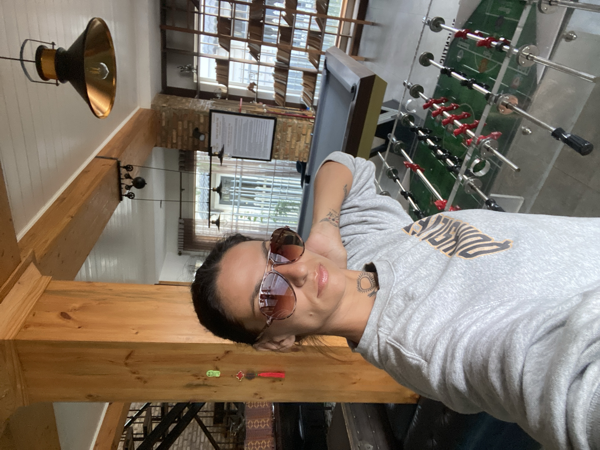 The image depicts a woman taking a selfie in what appears to be a recreational area of a building. She is wearing sunglasses and a grey sweatshirt. Behind her, a pool table and a foosball table are visible. The environment features wooden beams, brick walls, bookshelves filled with books, and hanging industrial-style light fixtures. The scene suggests a casual, perhaps indoor sports or lounge setting, possibly within a hotel or a private residence. The presence of a small green smiley-faced decoration and a red tassel ornament hanging on a wooden pillar hints at a possible decorative theme or cultural influence. The building's architecture and natural wood elements might suggest a location in Da Lat, Vietnam, a city known for its French colonial architecture and surrounding natural beauty.