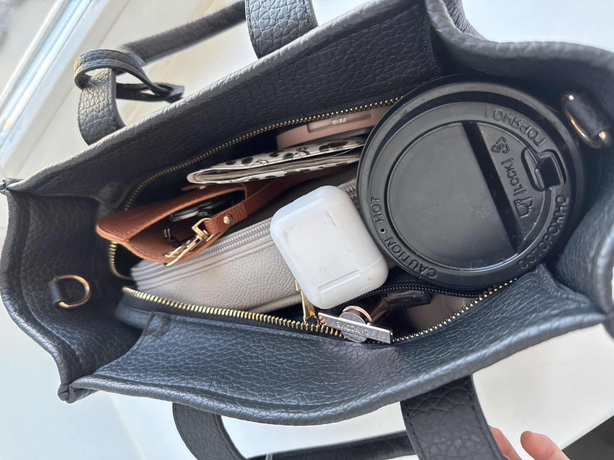 This high-angle, close-up shot captures the contents of an open, black textured leather bag, likely a tote or handbag, in Chaikovsky, Russia. The bag features a pebbled leather finish and prominent gold-toned zippers. The scene is brightly lit by diffused light, suggesting daytime, possibly near a window.
Inside the main compartment of the bag, various personal items are nestled together. A black plastic coffee cup lid, partially obscuring other items, is prominent on the right. Visible text on the lid includes "CAUTION," "HOT," a recycling symbol, and "LOCK," along with the word "TOP" written upside down.
Toward the center, a white Apple AirPods case, showing some signs of wear, rests on top of a light grey zippered pouch. To its left, a small, rich brown leather pouch or wallet with a gold zipper and clasp is partially visible. Further into the bag, a light pink compact or small case is tucked away, and a piece of patterned fabric, possibly a wallet or small planner, can be seen beneath it. A silver rectangular object, likely a keychain or a cosmetic item, bearing the embossed name "LANCOME," is visible near the AirPods case. The interior of the bag appears to be lined with black fabric. The image portrays the collection of daily essentials carried by an individual.
