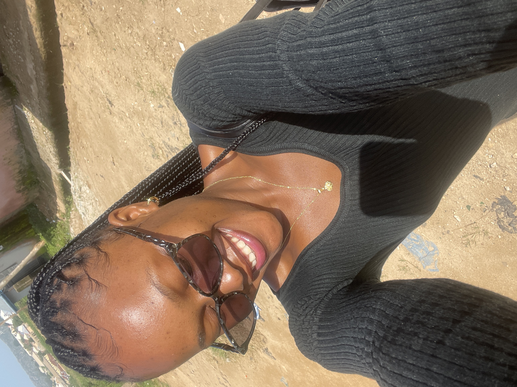 A young woman with braided hair and sunglasses is smiling for a selfie outdoors. She is wearing a black long-sleeved top and a gold necklace with a small pendant. The background appears to be a dirt path with some buildings and vegetation visible in the distance, suggesting an outdoor urban environment. The lighting indicates it is a sunny day. The scene suggests a casual, everyday moment.