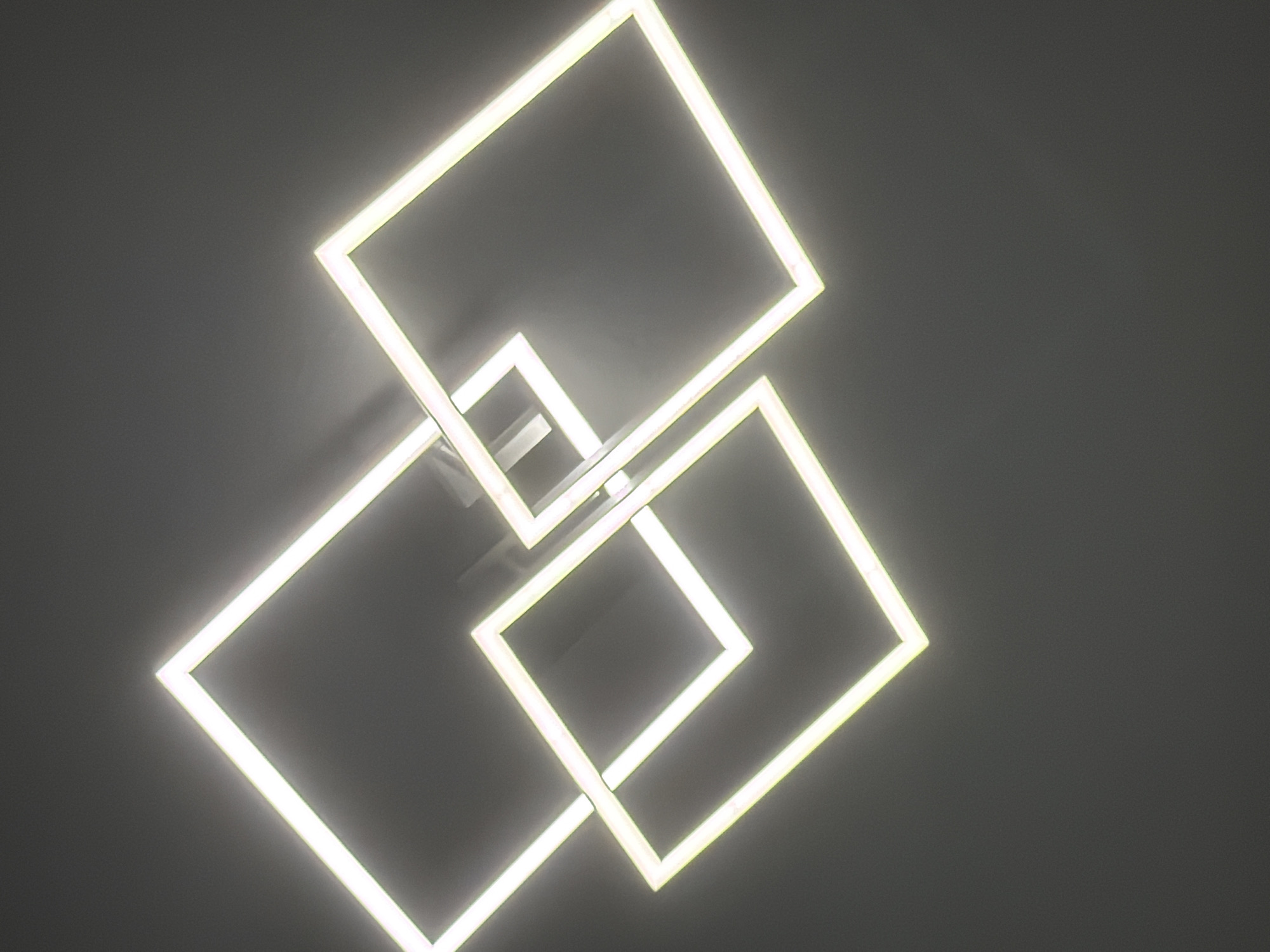 The image shows a modern, geometric ceiling light fixture with multiple interlocking square frames emitting a bright, cool white light against a dark gray ceiling. The fixture is suspended from the ceiling and appears to be made of metal with integrated LED lighting. The light creates a halo effect on the ceiling, and the metallic frames have a slight sheen. The composition is abstract, focusing on the interplay of light and geometric shapes. There are no people or other objects visible in the frame. The environment is indoors, likely a room with a dark ceiling. No specific location in Dorsten, Germany can be determined from the image alone.