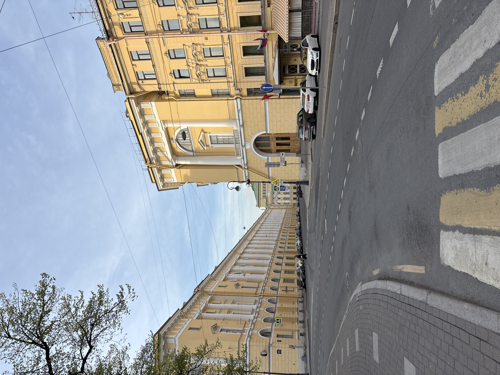 The image depicts a street scene in Saint Petersburg, Russia. The view is of a wide street with a crosswalk. On either side of the street stand tall, ornate, yellow buildings, typical of the city's architecture. Cars are parked along the right side of the road. Power lines stretch across the sky. A tree with green leaves is visible in the lower-left corner. The sky is light blue with scattered clouds.