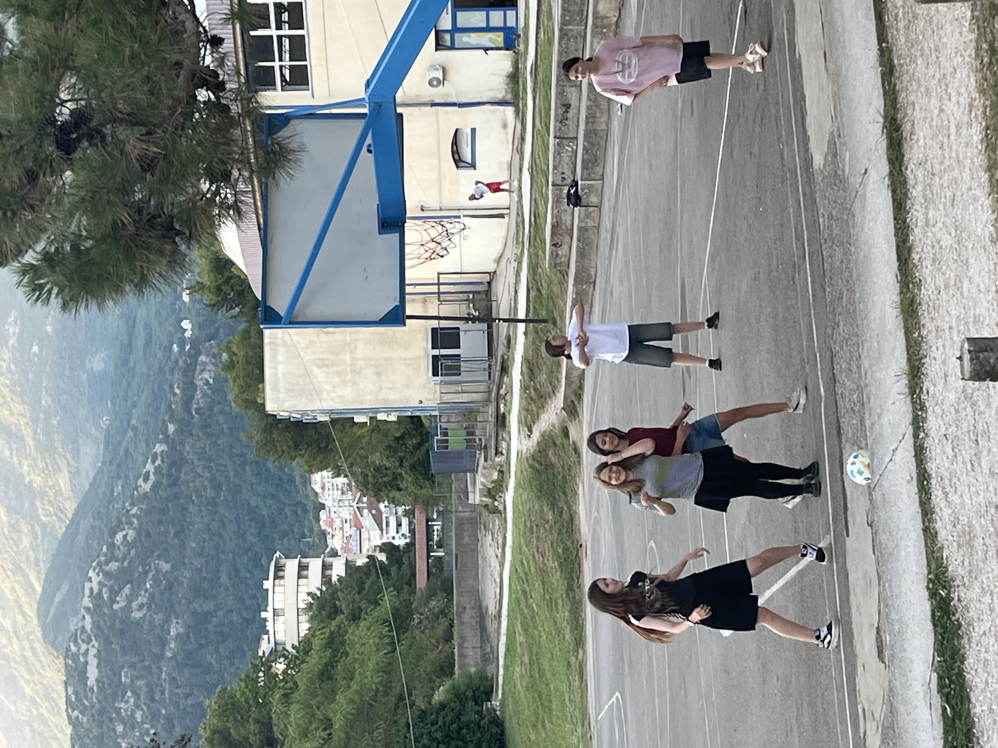 The image shows a group of five people standing on an outdoor basketball court next to a building with a basketball hoop. Behind the building is a view of mountainous terrain with buildings of Budva, Montenegro. One person wears a pink shirt and black shorts. One person wears a white shirt and gray shorts. Two girls are standing next to each other, one wearing a maroon shirt and jean shorts, and the other wearing a gray shirt and black skirt. A fifth person, another girl, is wearing a black dress and black-and-white shoes. A soccer ball is on the court near the group of three girls. The location appears to be near or in Budva, Montenegro, considering the buildings and the surrounding mountainous environment.