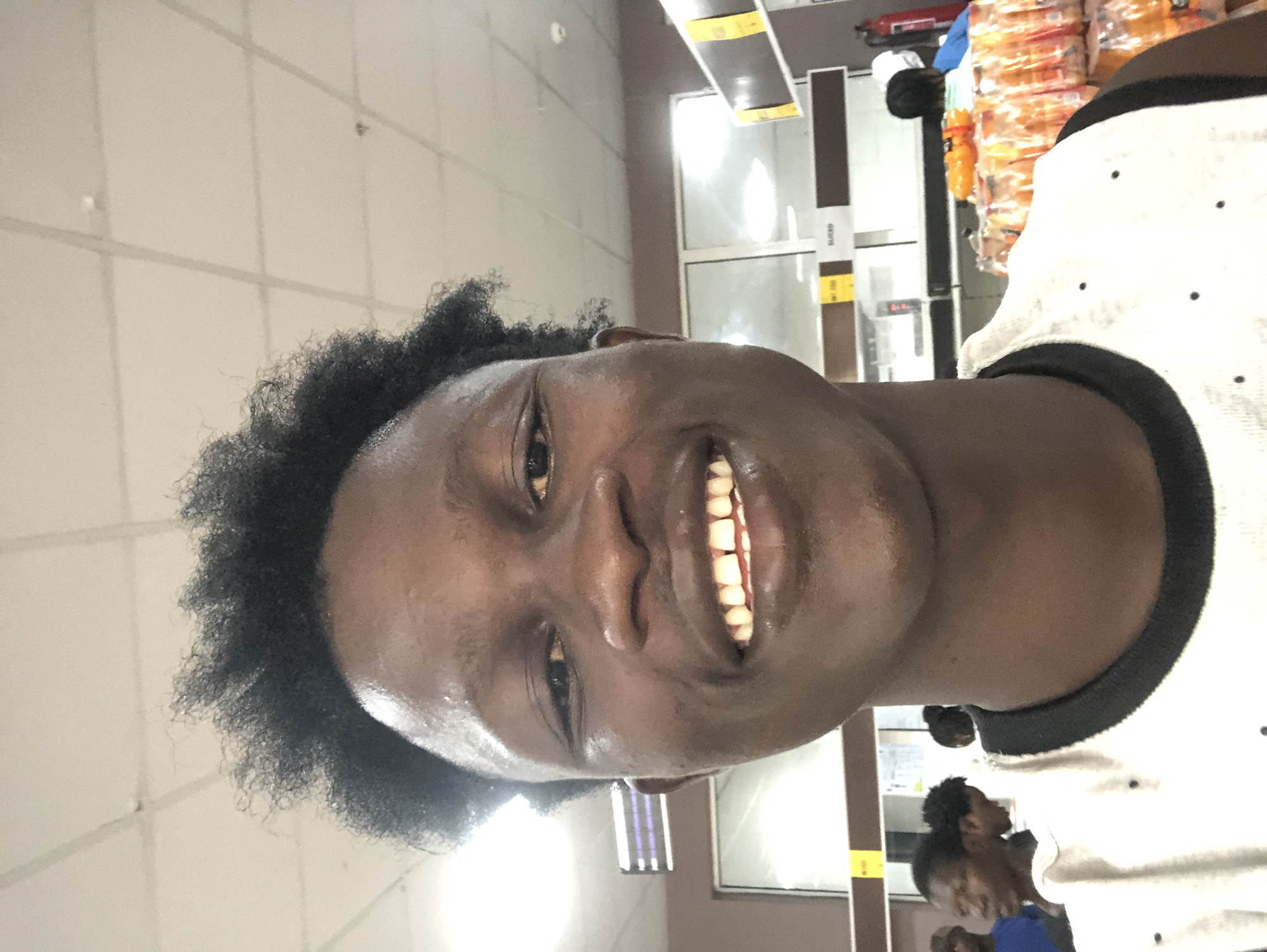 In Ado, Nigeria, a young Black male with dark, curly hair is taking a selfie in what appears to be a supermarket. He is smiling, showing his teeth. He is wearing a white tank top with a black trim. In the background, other shoppers are visible, as well as aisles stocked with products. The ceiling is a tiled drop ceiling with fluorescent lighting.