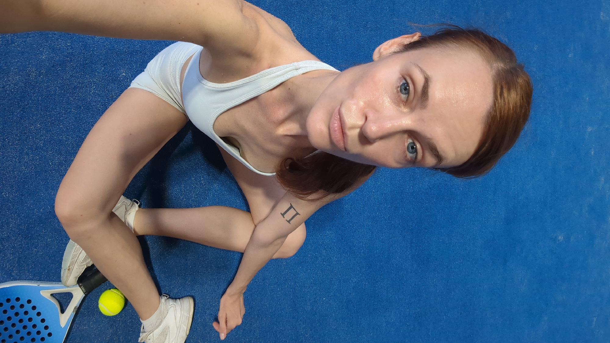 A woman with reddish-brown hair is sitting on a blue surface, possibly an indoor court. She is wearing a white athletic top and shorts, and white sneakers. She has a tattoo on her left forearm that appears to be the Roman numeral II. A tennis racket and a tennis ball are visible near her feet. The setting does not provide any specific visual cues to place it in North Kuta, Indonesia.