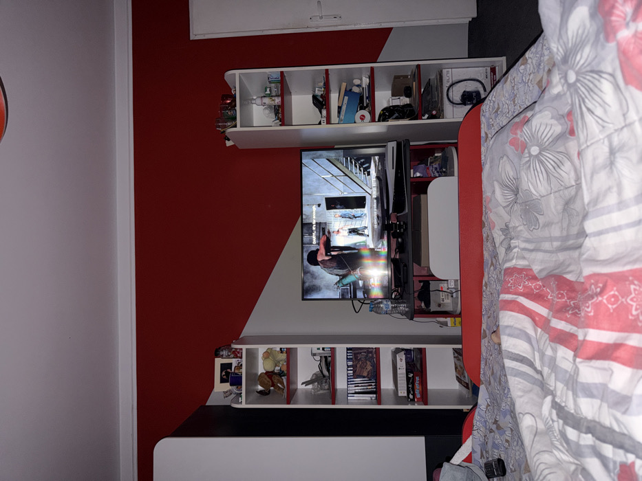 The image captures an interior room in Opanets, Bulgaria, viewed from a low perspective, consistent with being observed from a bed. The room features a white ceiling and walls painted with a striking diagonal pattern, primarily red in the upper section transitioning to white below. A large flat-screen television is centrally positioned on a dark grey entertainment stand. The screen displays a scene from what appears to be a video game or television show, featuring a male character in the foreground aiming a crossbow-like weapon, with two other male figures in the background within an industrial or institutional setting with metal stairs. Text on the screen includes "DARYL: It's far enough" and "натиснете продължи" (Bulgarian for 'press continue').
Below the television, a white PlayStation 5 console rests on the entertainment stand, alongside a plastic water bottle with a blue cap and various electronic cables. The entertainment setup is flanked by two tall, narrow white shelving units with red shelf backings. The left unit contains items such as a framed photograph, two stuffed animals, multiple video game cases, and books. The right unit holds assorted household objects, including drinking glasses, bottles, and storage containers.
In the immediate foreground, a bed with a patterned duvet featuring white, grey, and red floral and striped designs is visible. A red headboard or footboard extends beyond the duvet. A remote control is partially visible on the bed to the left. A white door with a silver handle is positioned on the far right side of the room, and dark flooring is discernible adjacent to it. No individuals are present in the frame.