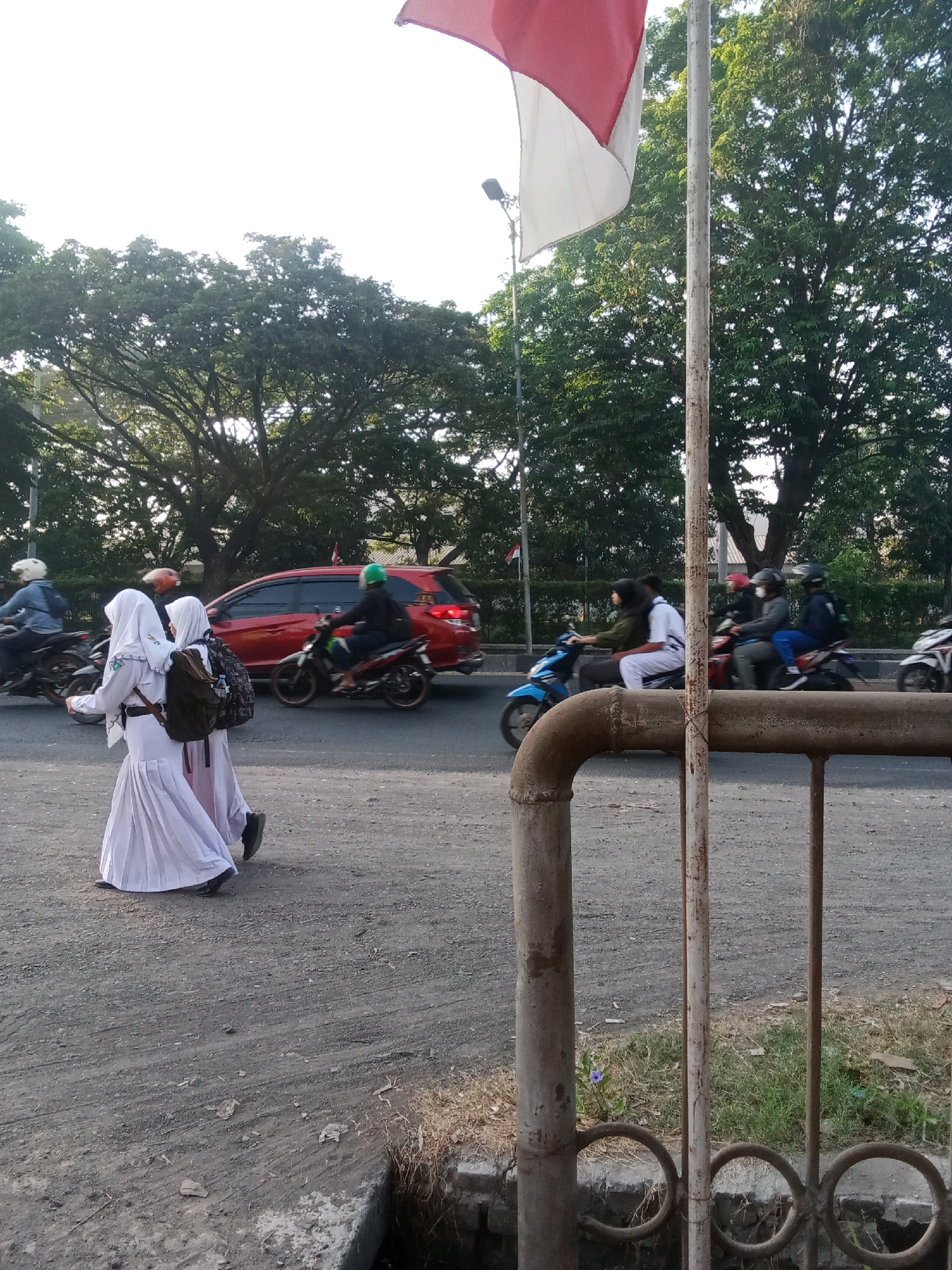 In the city of Keboharan, Indonesia, two young individuals wearing white school uniforms and headscarves walk on a gravel-laden road. They are seen from behind, with one carrying a backpack. The road appears to be a main thoroughfare, with several motorcycles and a red car passing by. In the background, lush green trees line the street, and a partially unfurled Indonesian flag is visible. The scene captures a moment of everyday life on a public road, with elements of transportation and education present.