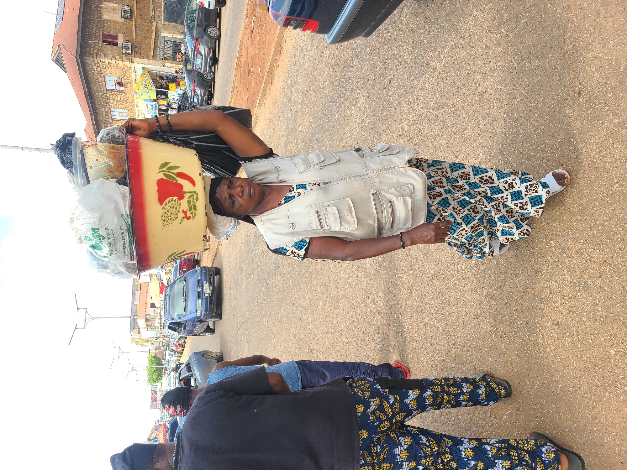 A woman balances a large yellow basin filled with various items on her head. She wears a light-colored vest over a patterned blue and white dress and a headscarf. Her left arm is raised, supporting the basin. In the foreground, two men are visible. The man closer to the camera wears a black t-shirt and patterned pants, with a black head covering. The man behind him wears a blue t-shirt and dark pants. The scene appears to be a street with vehicles, including a blue car and a dark-colored car, parked and driving. Buildings line the background. The lighting suggests it is daytime, and the weather seems overcast. There are no visible signs with legible text. The woman's expression is neutral as she carries her load.