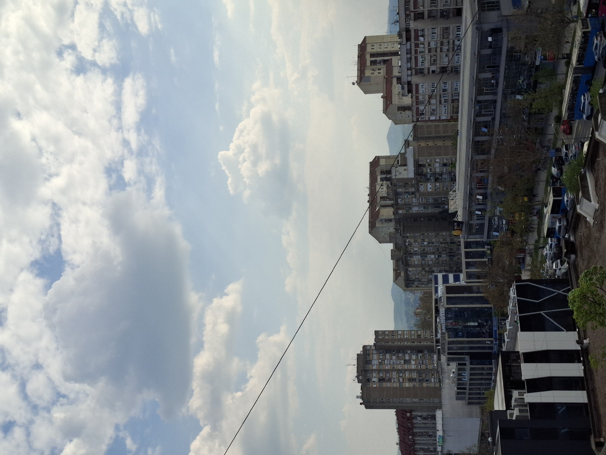This image depicts an urban landscape in Novi Sad, Serbia, viewed during daytime under a partially cloudy sky. The perspective is from an elevated position, looking across a dense area of buildings and streets.
The upper-left portion of the frame is dominated by a bright blue sky filled with numerous white, cumulus clouds, indicating fair weather. A dark power line or cable spans diagonally across the sky and cityscape, running from the upper-left to the lower-right of the image.
The right and lower sections of the image showcase a mix of architectural styles. Many buildings are multi-story residential blocks, characterized by their numerous windows and balconies, suggestive of post-war or socialist-era construction. Interspersed among these are more contemporary commercial buildings, some with modern glass facades and geometric designs. One prominent modern building features a blue sign on its facade with the text "AM HOTEL" clearly visible, and "PARKING" below it. Another yellow sign, partially visible on a different building, appears to read "BAK...".
At ground level, streets or parking areas are visible, populated with numerous parked cars. Scattered trees, some with green foliage and others with bare branches, add natural elements to the urban setting. In the distant background, hazy outlines of hills or mountains can be seen on the horizon, blending with the sky. No specific human activity or event is distinctly captured, but the scene reflects typical daily life in a bustling city. The bright lighting suggests it is mid-day to afternoon.
