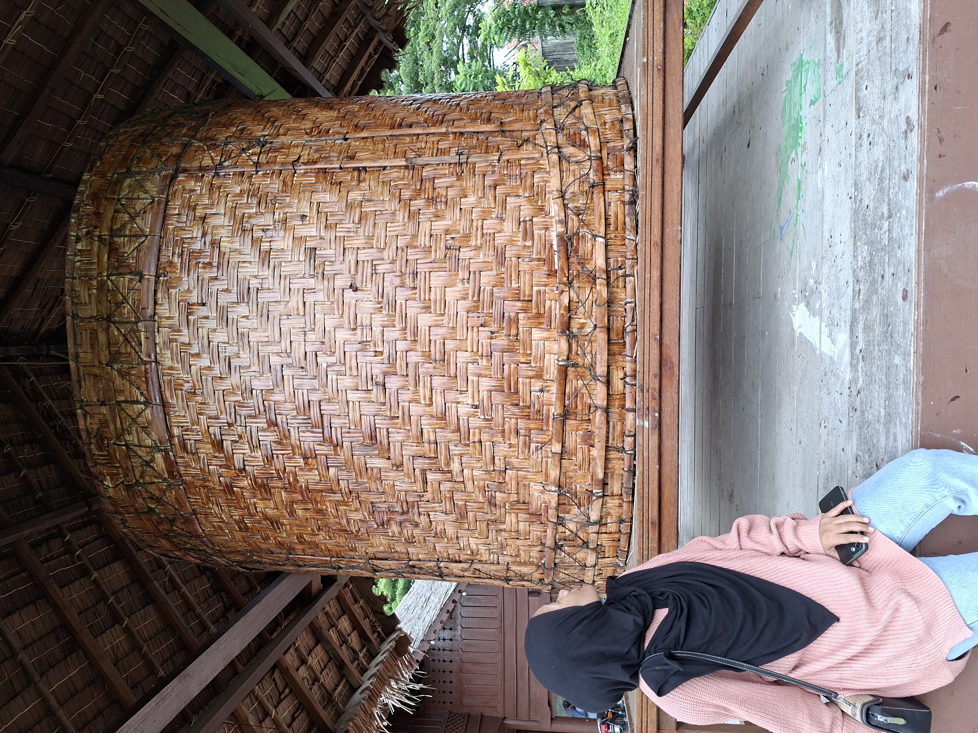 A horizontal, medium shot captures a woman in Lhokseumawe, Indonesia, seated next to a large, woven basket. The woman wears a light pink sweater, light blue jeans, and a black headscarf. She holds a black smartphone in her left hand, and a black purse strap crosses her torso. She is seated on a brown wooden platform.
The woven basket is the dominant feature of the image, taking up a significant portion of the frame. It is made of light brown woven material and held together with dark-colored ropes or bindings. The basket is positioned inside a structure with a thatched roof, visible in the upper left corner of the frame.
The background includes trees and greenery, indicating an outdoor setting. A partially visible building with brown wooden walls and traditional carvings is also present. The overall environment suggests a cultural or historical site in Lhokseumawe.