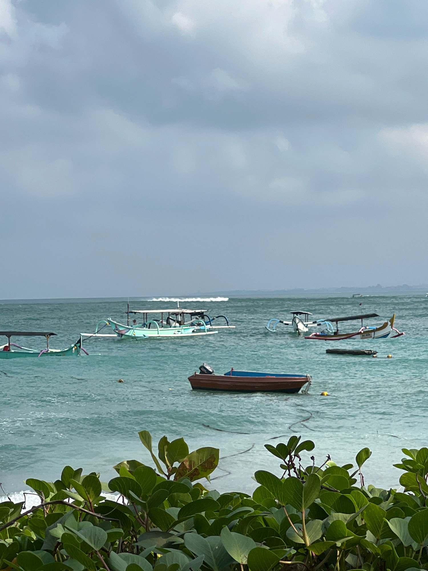 This is a medium shot, taken from a low angle, of a coastal scene in Kuta, Indonesia. The foreground is dominated by lush green foliage, with large, heart-shaped leaves and thick vines. Behind the foliage, a calm, turquoise sea stretches out to the horizon. Several traditional Balinese outrigger boats are scattered across the water. Two boats, painted a light turquoise, are anchored closer to the shore, each with a dark canopy providing shade. Further out, a larger boat with a white canopy and colorful decorative elements on its hull is also visible. In the distance, a few more boats can be seen, and a faint line of white waves suggests a break in the distance. The sky is overcast with a layer of gray clouds, creating a soft, diffused light. The overall impression is one of tranquility and a typical day on the coast of Kuta.