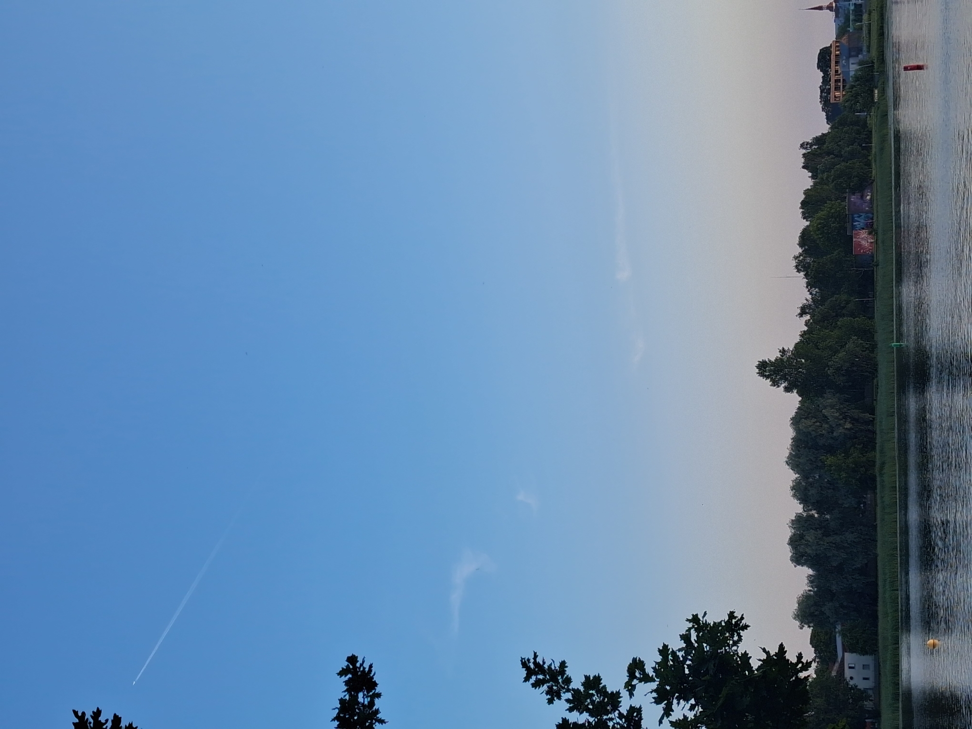 A wide shot captures a serene lake in Pärnu, Estonia, under a clear, light blue sky with a subtle gradient towards the horizon. A faint contrail from an airplane stretches across the upper left quadrant of the sky. The left foreground is dominated by the dark, silhouetted branches of a leafy tree. The middle ground features a dense line of green trees bordering the lake, interspersed with structures including a white building on the left and a building with colorful graffiti on the right. A spire of a distant church is visible on the far right. The water surface of the lake is calm, reflecting the sky and trees with subtle ripples. Several buoys, one yellow in the left-middle of the lake and a red one on the right, mark the water. The overall impression is one of tranquility at dusk or dawn.